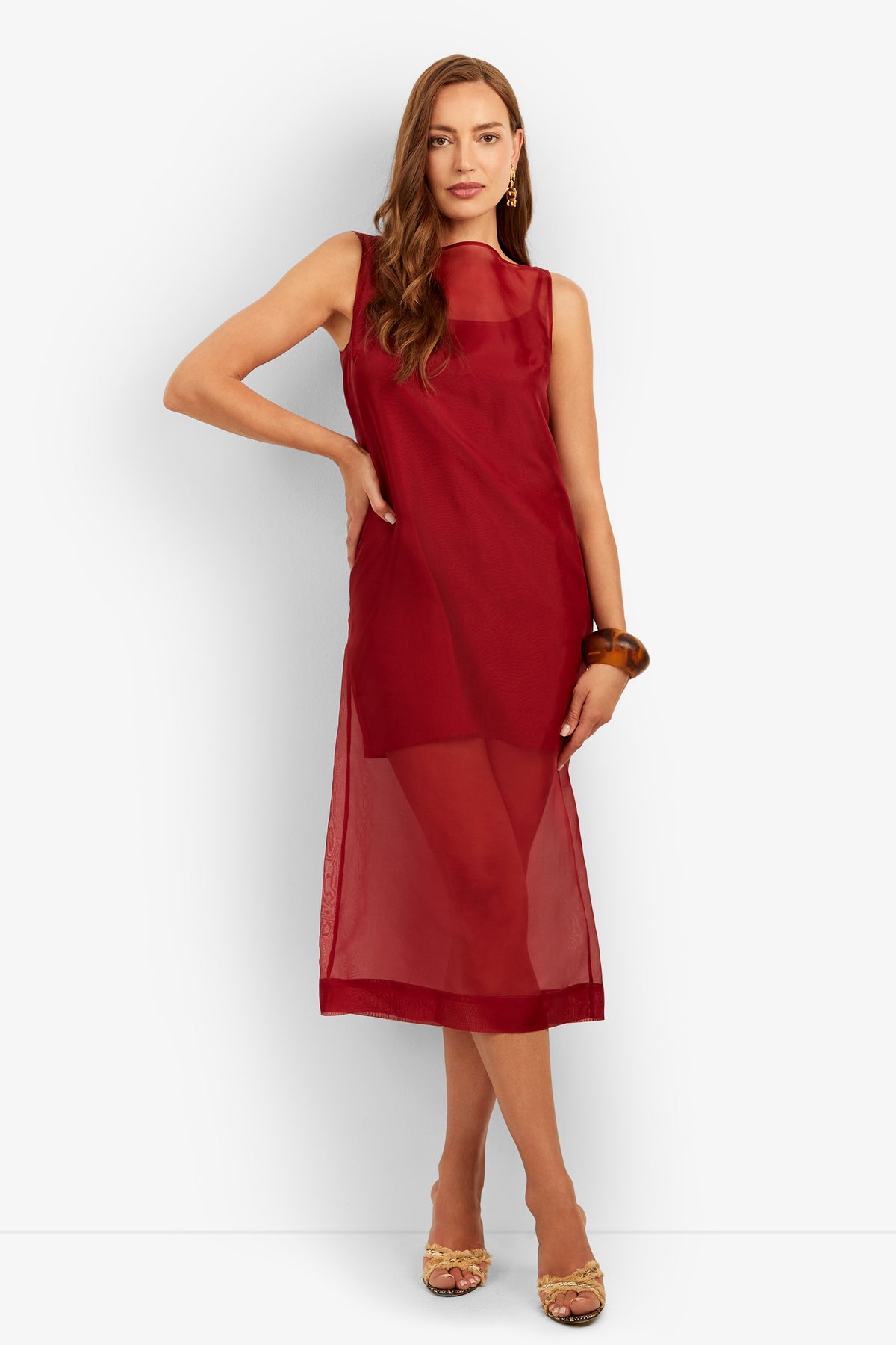 A woman wearing the Roselle Dress—a sleeveless, sheer red midi dress—stands with one hand on her hip. She has long wavy hair, gold earrings, a brown bangle, and beige sandals against a plain white background.