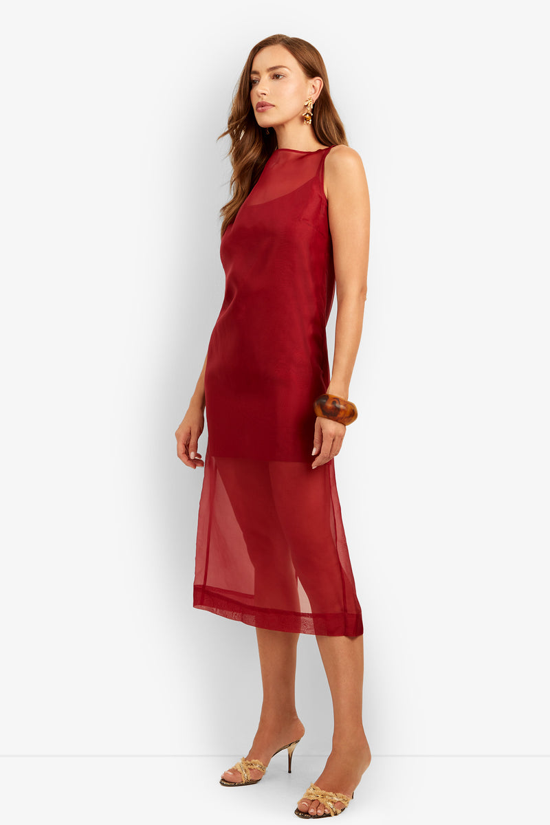 A woman models the Roselle Dress—a sleeveless, knee-length sheer red dress with a high neckline. She pairs it with long hair, gold earrings, a brown bangle, and strappy heels while standing against a plain white background.