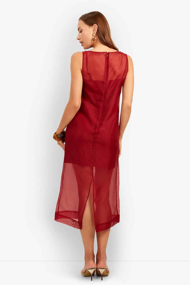 A woman stands facing away, wearing the Roselle Dress—a sleeveless, sheer red midi dress featuring a back slit. She has straight brown hair, gold earrings, a bracelet, and beige heels. The background is plain white.