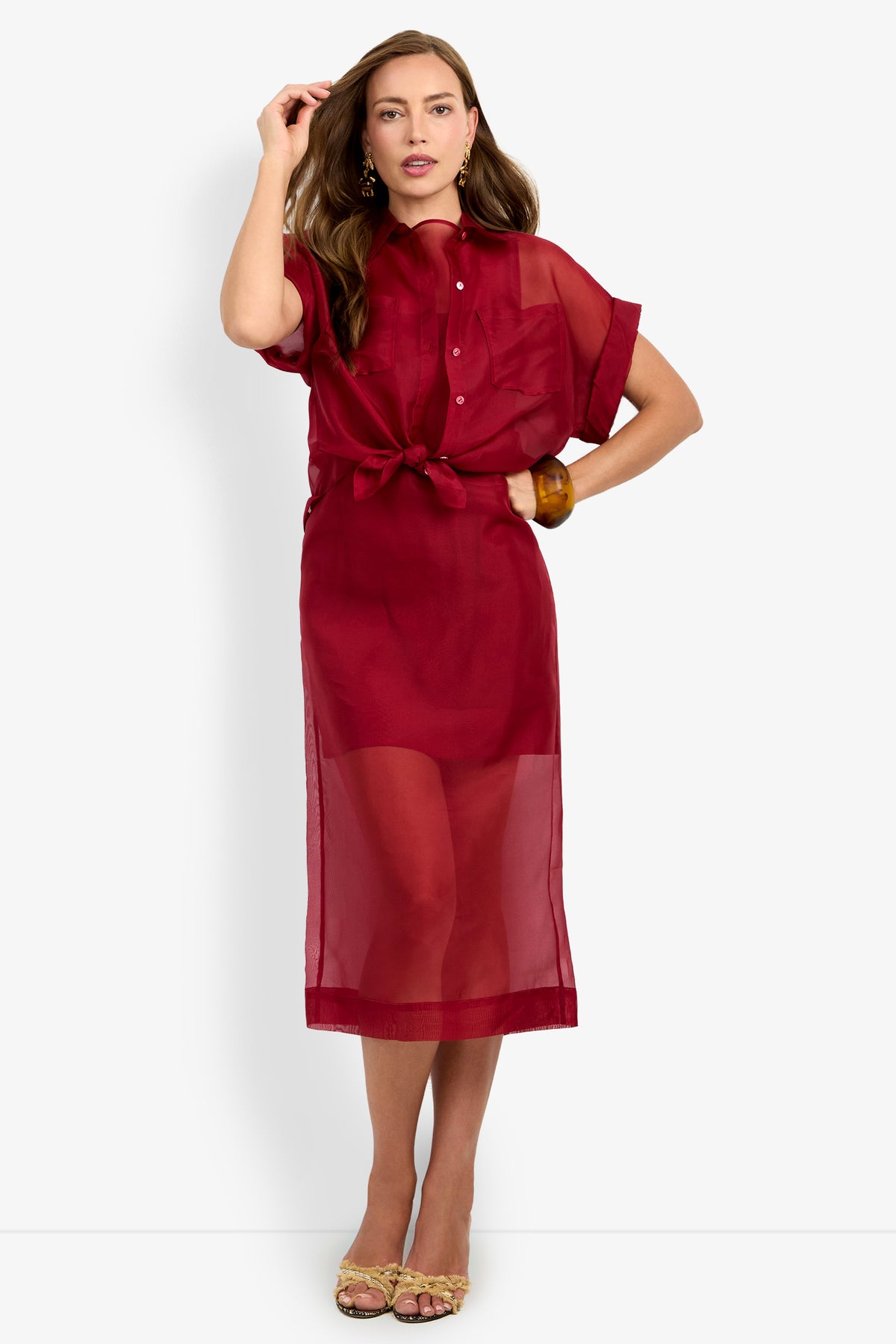 Model wearing the COSTANZO TOP, a sheer, short-sleeve, button-up red blouse with a front knot, paired with a matching red skirt underneath, gold earrings, and strappy sandals, standing against a plain white background.