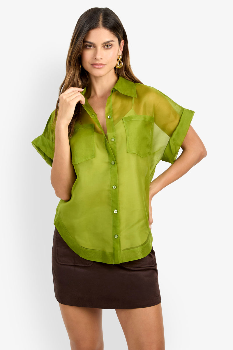 A woman with long brown hair wears the COSTANZO TOP, a sheer green short-sleeve button-up shirt with front pockets, styled with a short dark brown skirt against a plain white background.