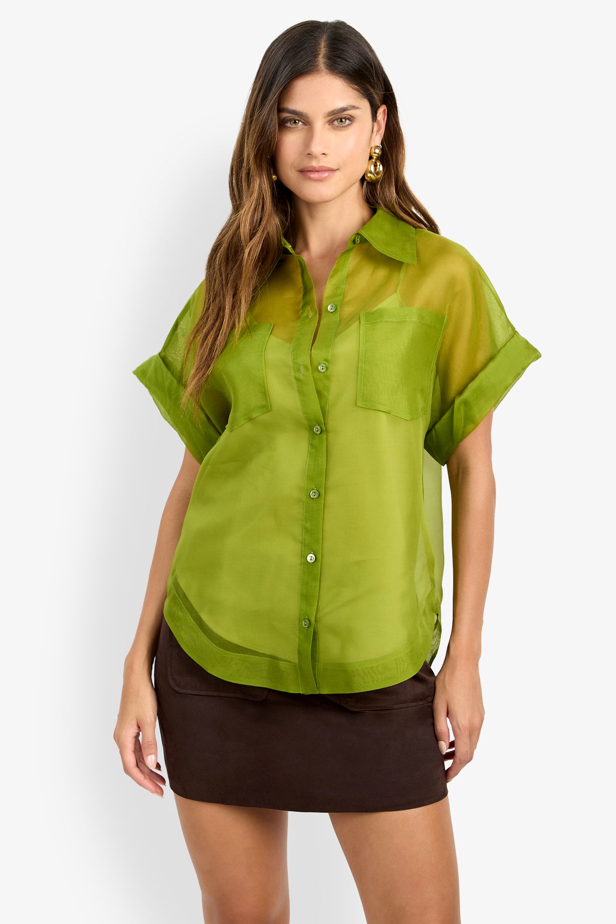 Woman wearing the COSTANZO TOP—a sheer green short-sleeve button-up shirt with a chest pocket—paired with a short dark brown skirt, standing against a plain light background.