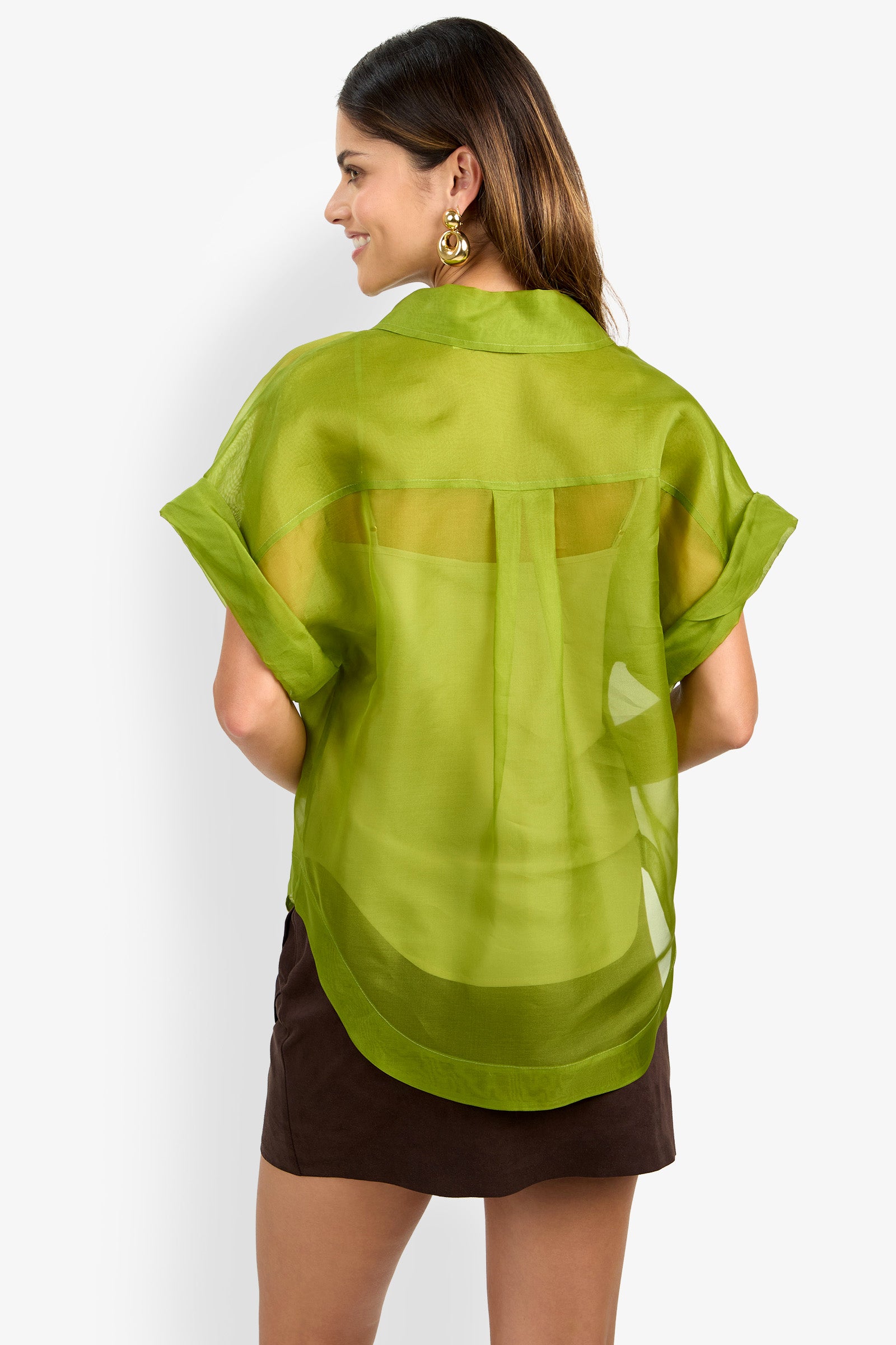 Woman smiling and facing away, wearing the COSTANZO TOP—a sheer green short-sleeve blouse with rolled sleeves and a back pleat—paired with a dark brown skirt. She has long hair and accessorizes with large gold earrings.