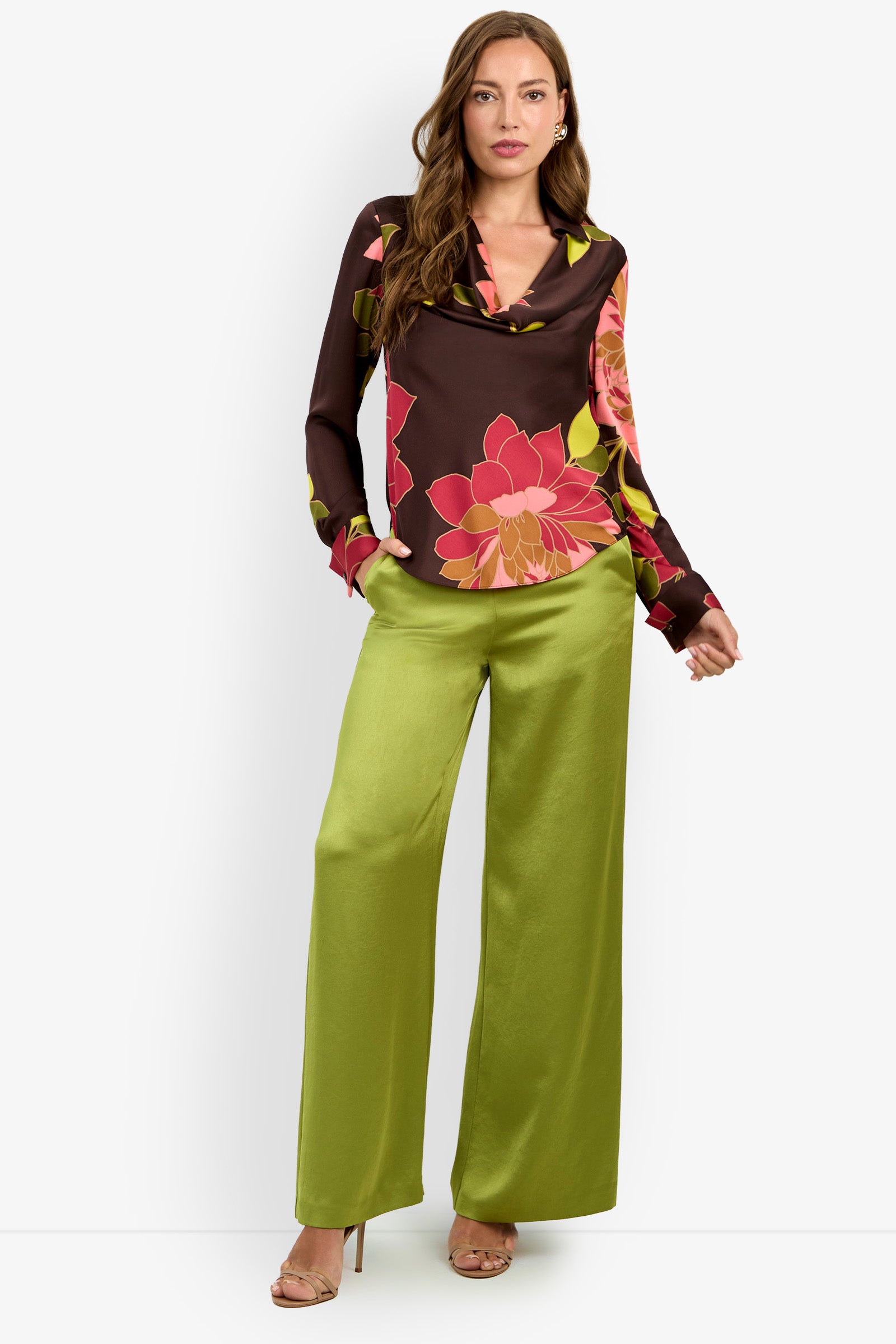 A woman models the Mistic Top, a dark floral blouse adorned with pink and yellow flowers, paired with loose, shiny lime green pants and beige high-heeled sandals. She stands against a plain white background.