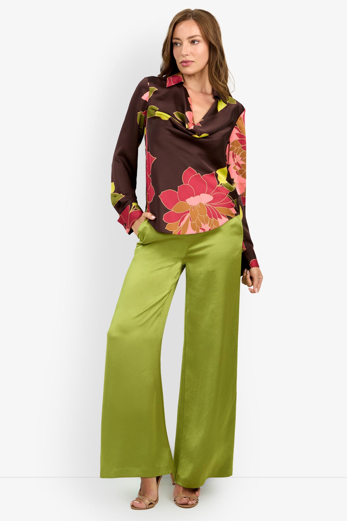 Woman wearing the Mistic Top, a brown blouse with large pink and yellow floral patterns, paired with wide-leg satin green pants and beige open-toe heels, standing against a plain light background.
