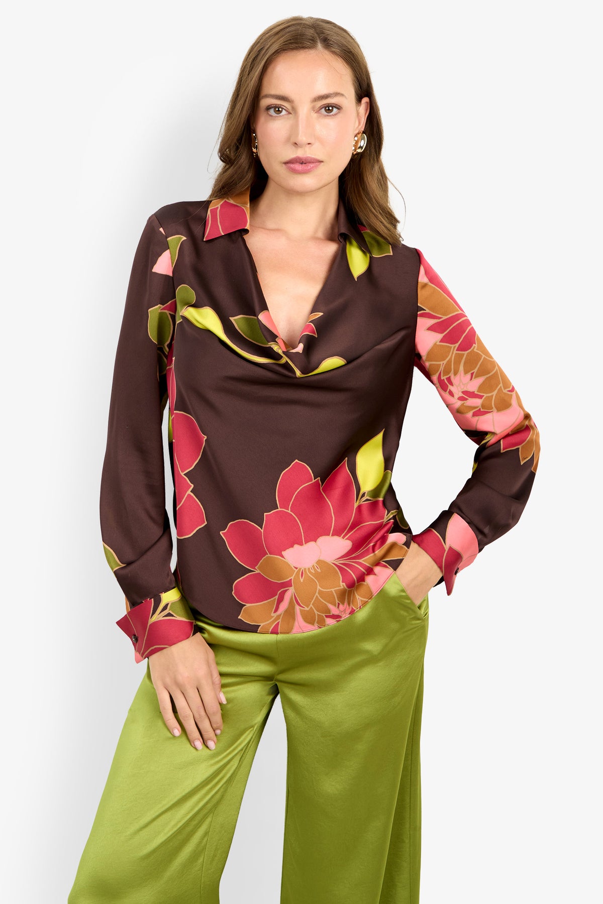 A woman wearing the Mistic Top, a brown blouse with pink and red floral print, paired with light green satin pants, stands with one hand in her pocket against a plain white background.