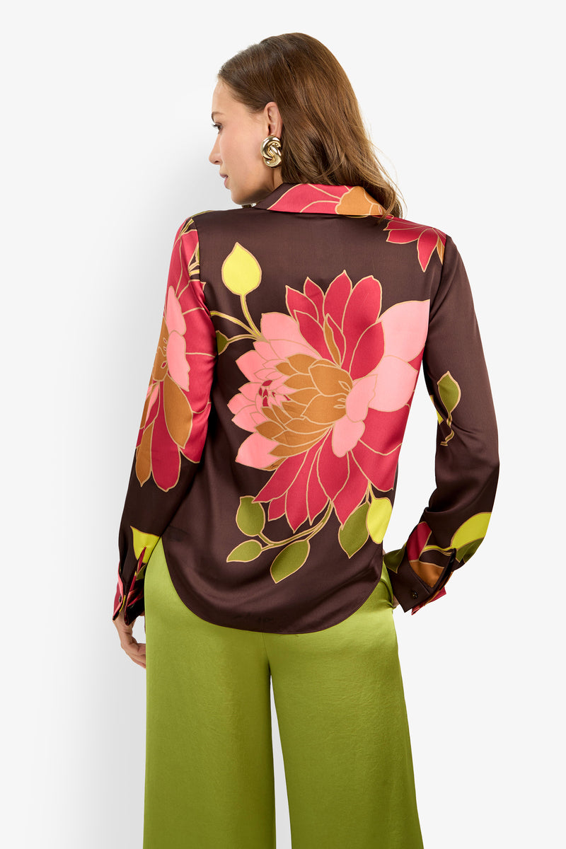 Woman facing away, wearing the Mistic Top—a brown blouse featuring a large pink and red floral print—paired with light green wide-leg pants. She has long brown hair and round earrings. The background is plain white.