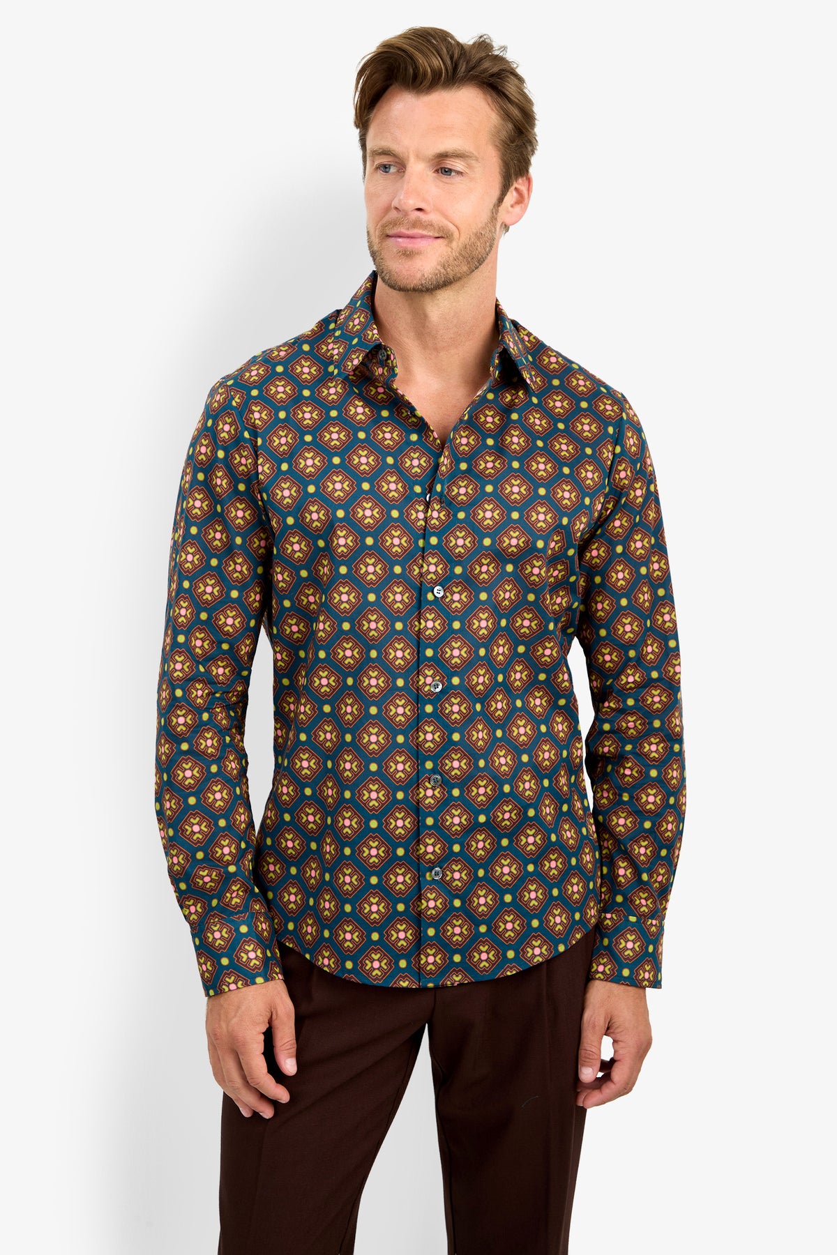 A man wearing the Aaron Shirt—a long-sleeve, button-up shirt featuring a colorful geometric pattern—stands against a plain white background, smiling as he gazes slightly to the side.