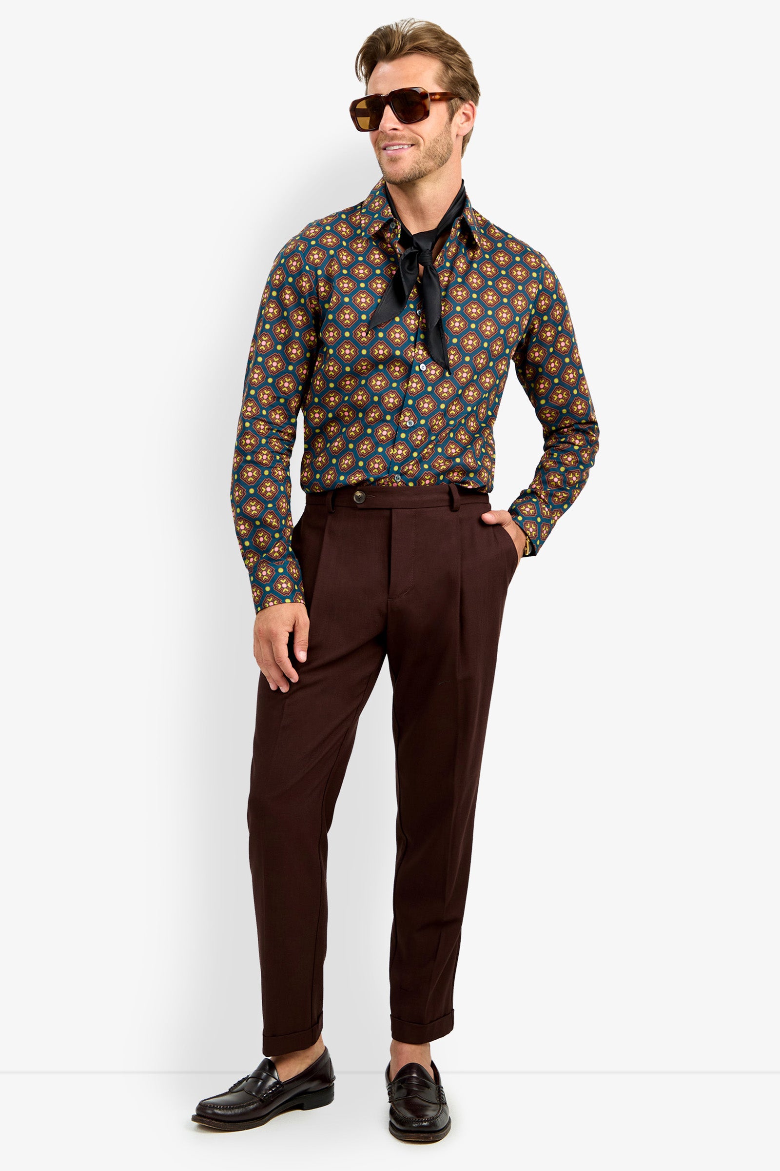 A man wearing the Aaron Shirt, styled with sunglasses, a neck scarf, brown high-waisted trousers, and black loafers, poses with one hand in his pocket against a white background.