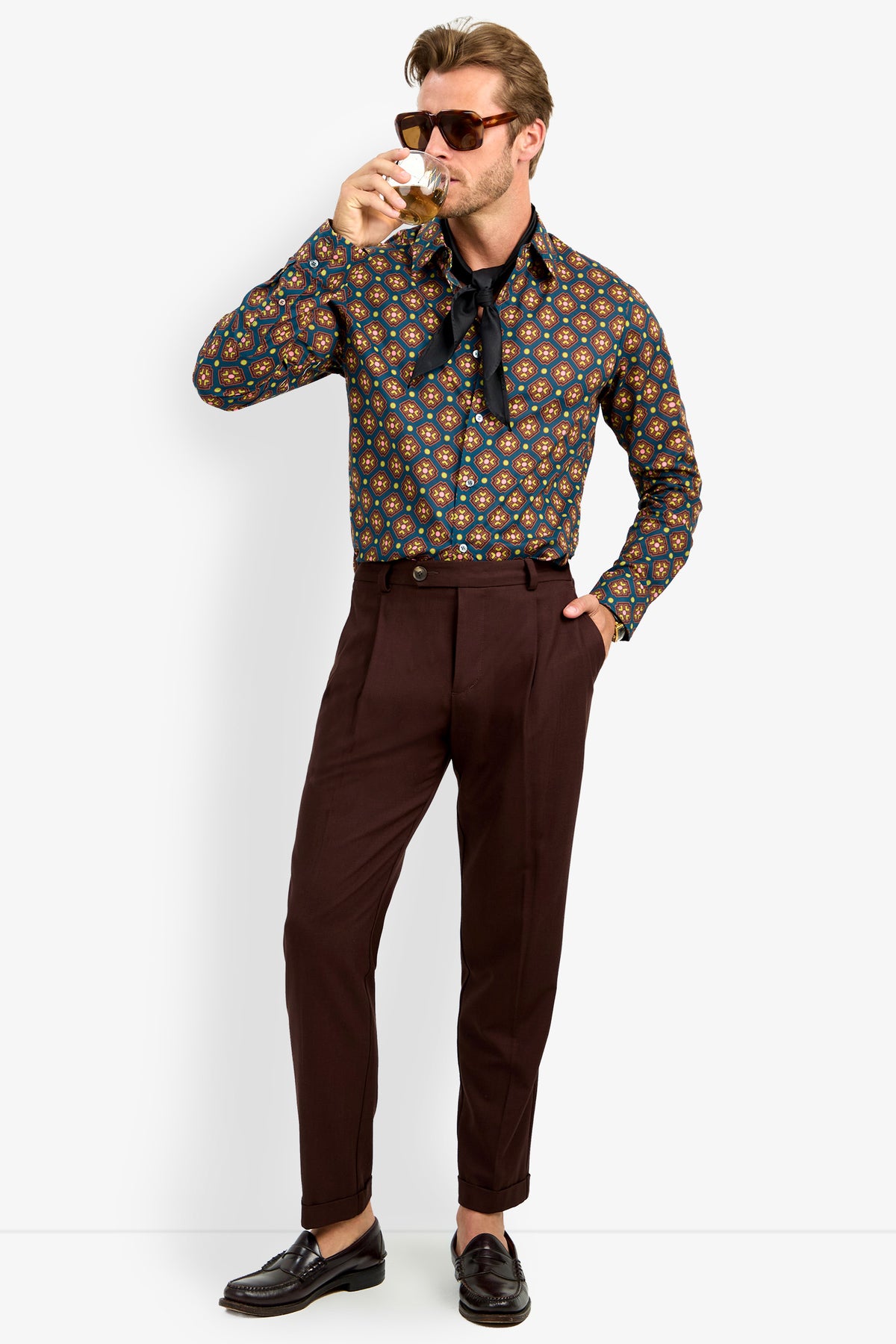 A man wearing the Aaron Shirt, sunglasses, dark brown pants, and loafers sips a drink with one hand in his pocket and a loosely tied scarf around his neck.