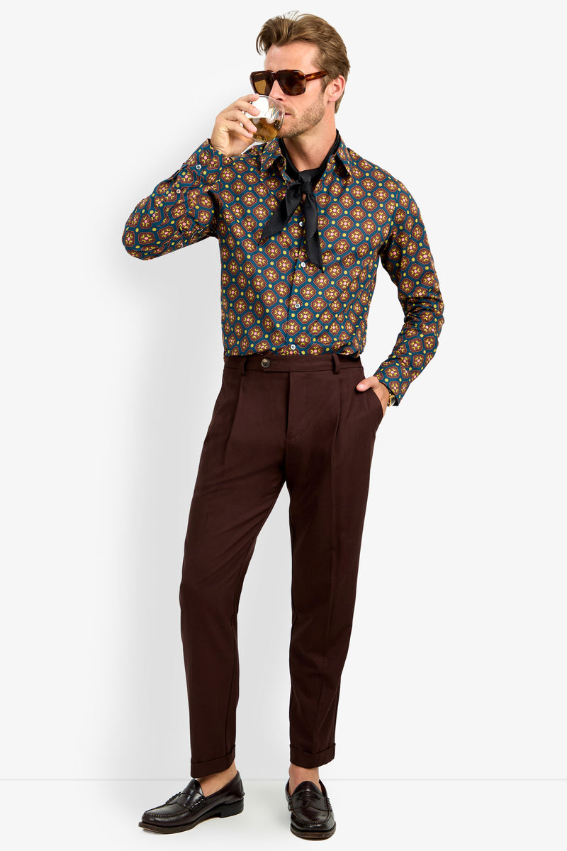 A man wearing the Aaron Shirt, sunglasses, dark brown pants, and loafers sips a drink with one hand in his pocket and a loosely tied scarf around his neck.