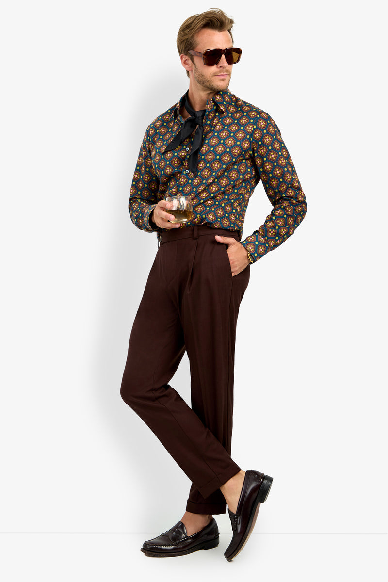 A man in sunglasses, wearing the Aaron Shirt with brown trousers and loafers, holds a drink while standing casually against a white background.
