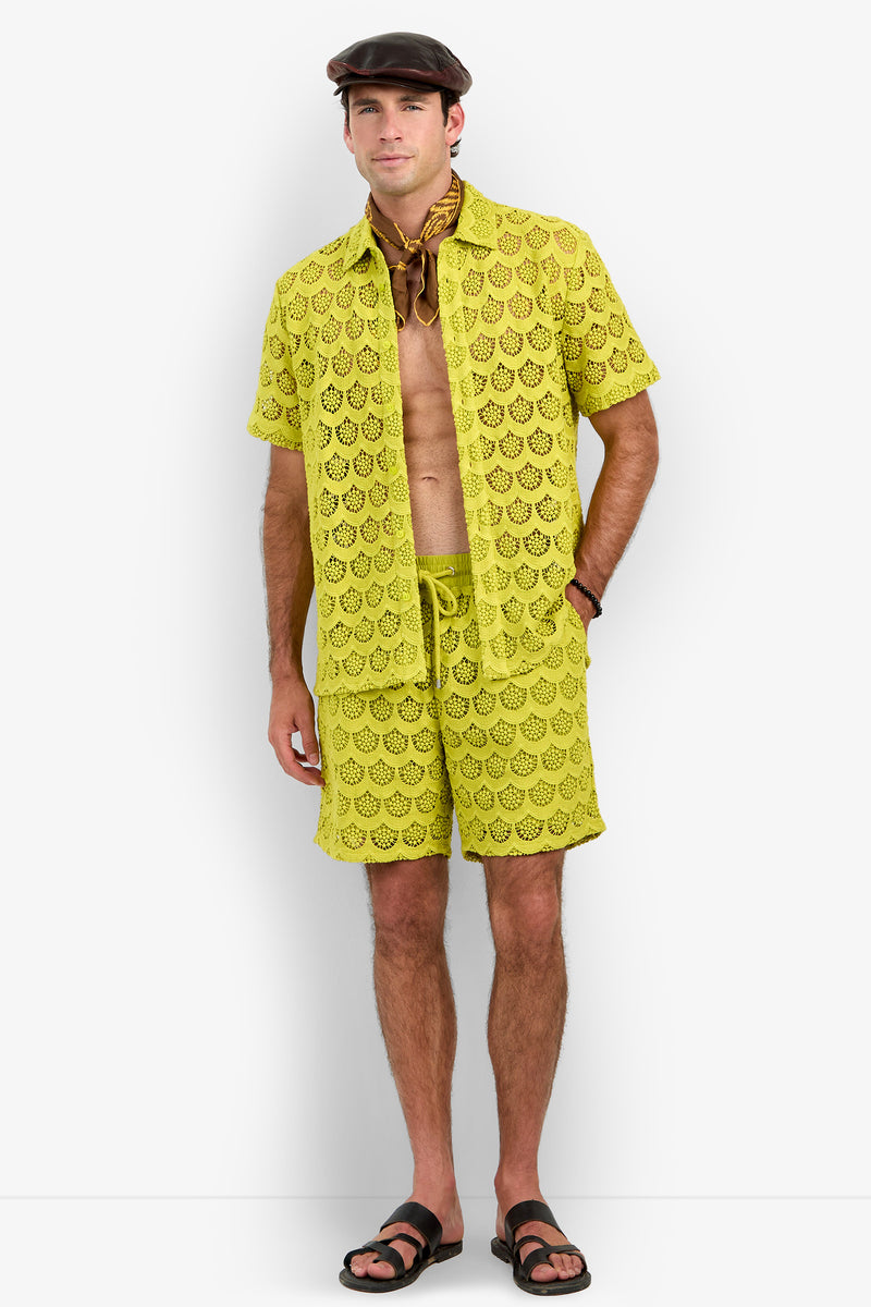 Wearing the Branson Shirt in yellow lace paired with matching shorts, a man completes his look with a brown flat cap, a brown neck scarf, black sandals, and a watch as he stands against a plain white background.