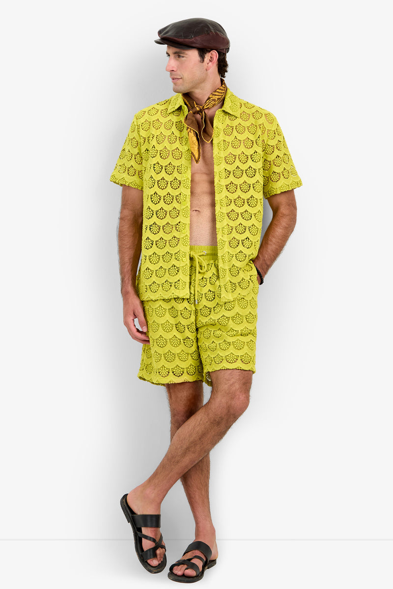 A man wears the Branson Shirt—a yellow lace short-sleeve shirt—paired with matching shorts, a brown flat cap, a brown neck scarf, and black sandals. He stands with one leg crossed over the other, looking to his left.