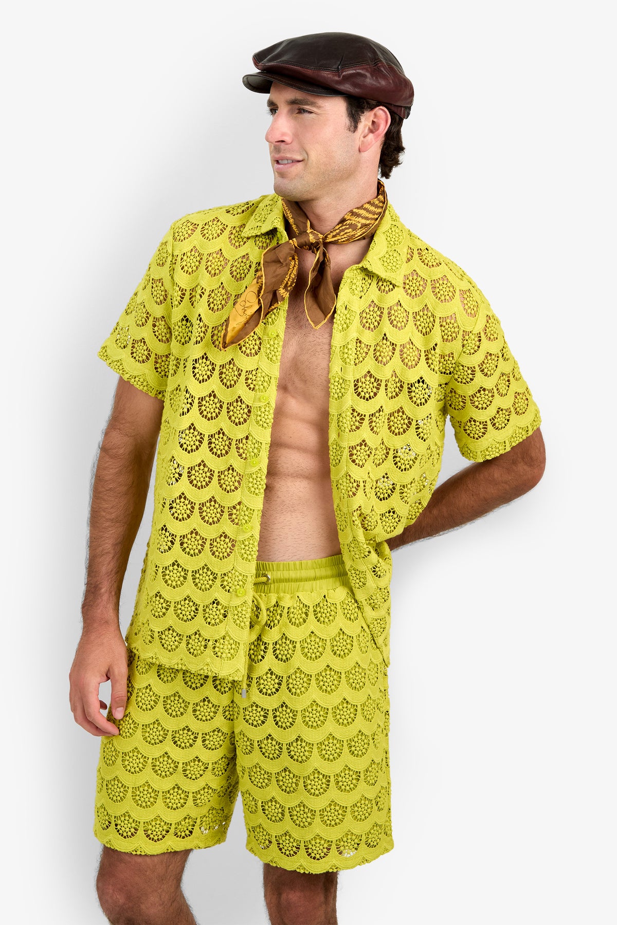 A man wearing the Branson Shirt, a yellow crocheted top paired with matching shorts, along with a brown leather cap and a gold patterned scarf around his neck, stands with one hand on his hip, looking to the side.