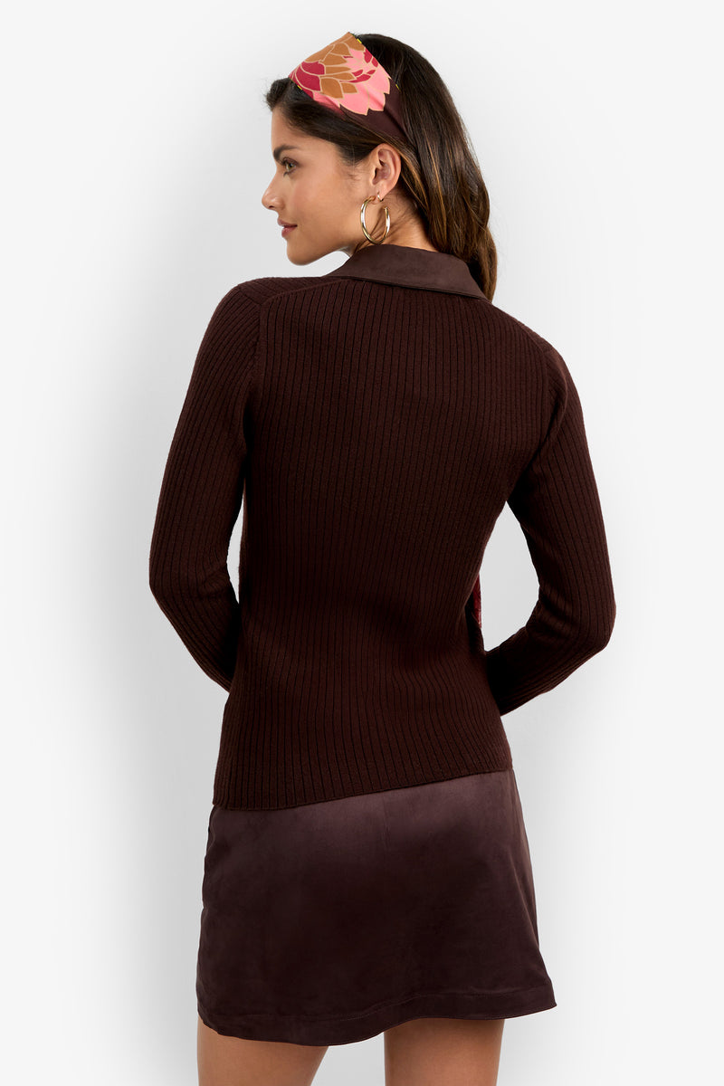 A woman with long brown hair, facing away, wears the Rita Long Sleeve Polo—a ribbed, dark brown top—paired with a mauve skirt. She accessorizes with large hoop earrings and a pink and tan geometric hairband.