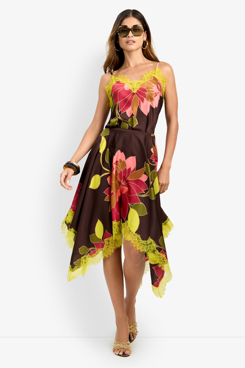 A woman wearing sunglasses and sandals holds one side of the Johari Skirt—a sleeveless, brown handkerchief-hem skirt featuring bold red and yellow floral prints with yellow lace trim. She has long hair.