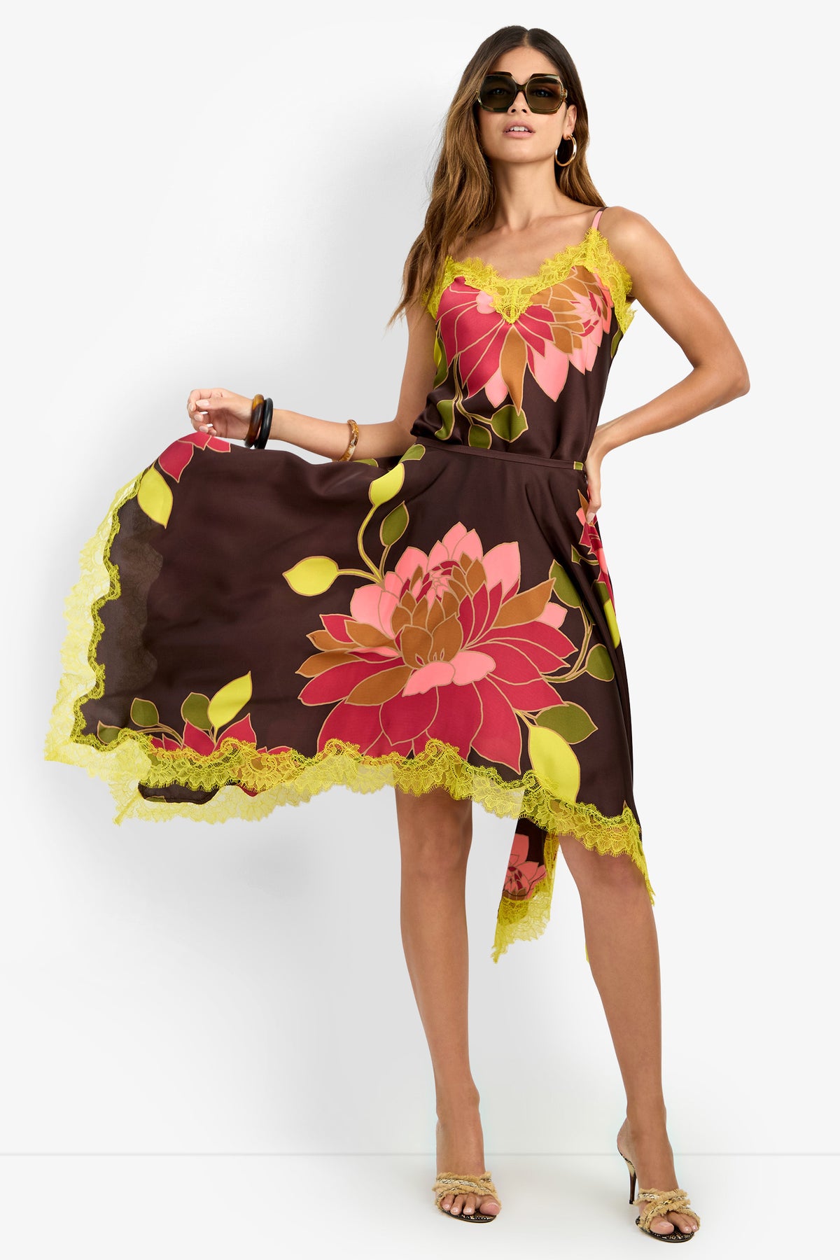 A woman wearing sunglasses and beige heels showcases the Johari Skirt, a brown asymmetrical piece adorned with large pink and yellow floral prints and finished with yellow lace trim, holding out one side of the skirt.