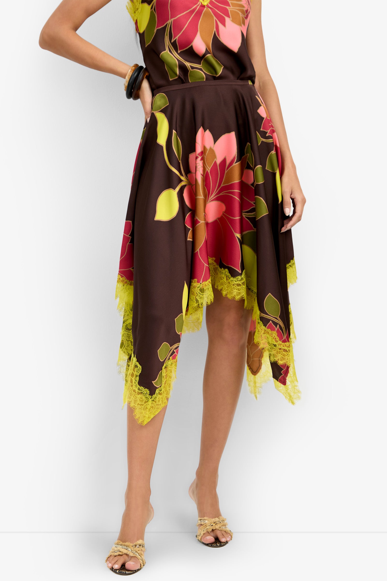 A woman stands against a white background wearing the Johari Skirt, a brown asymmetrical design with large pink and green floral prints and yellow lace trim, paired with tan open-toe heels.