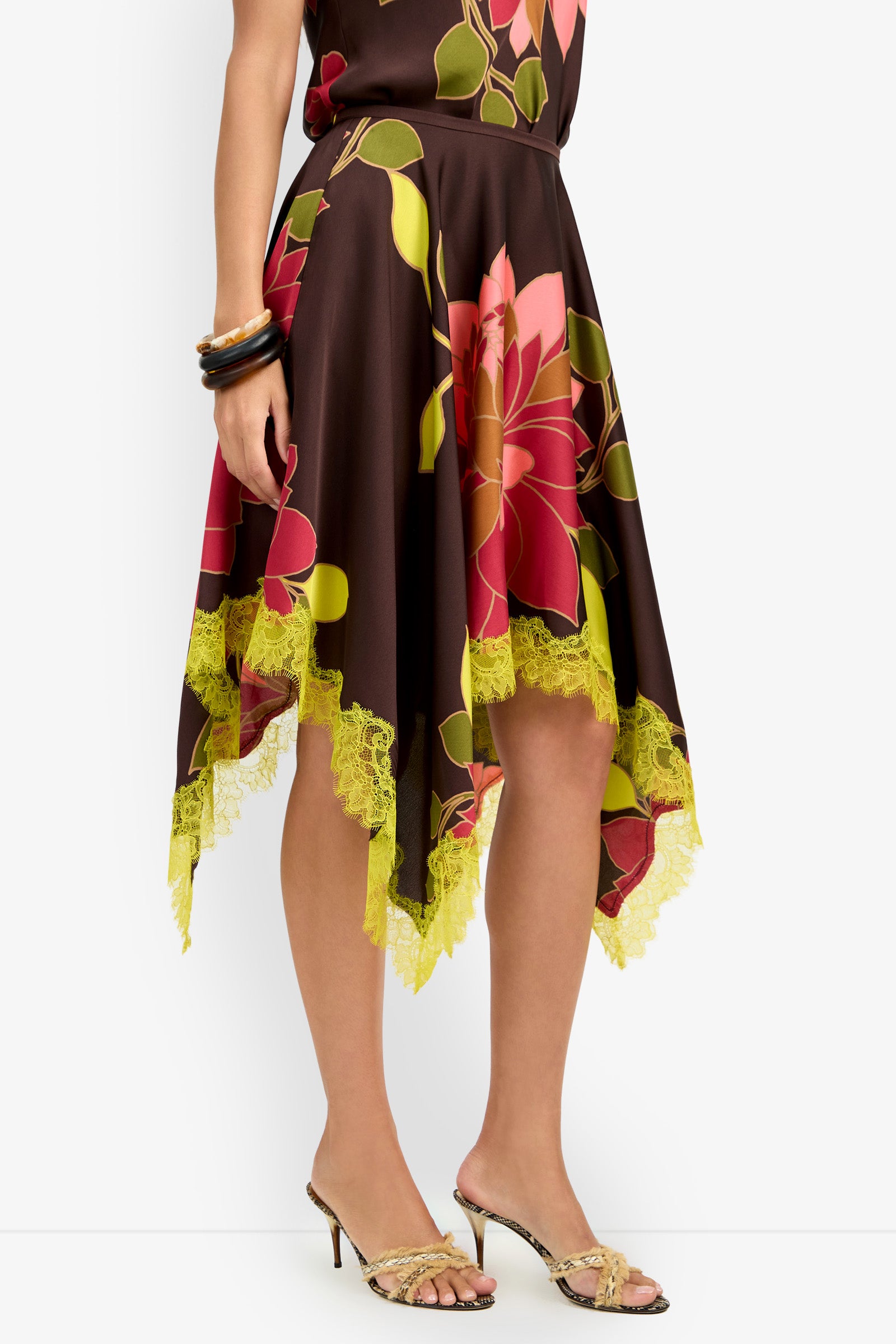 A woman wears the Johari Skirt, a brown piece with a handkerchief hem adorned with bold pink and green floral prints and yellow lace trim, paired with a matching top, gold heels, and black bangles.