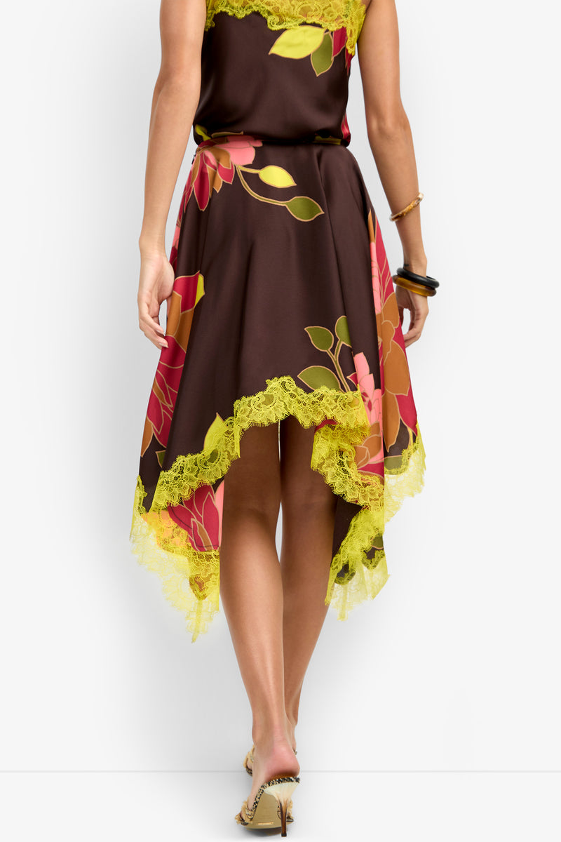 A woman stands with her back to the camera, showcasing the Johari Skirt—a brown floral piece with yellow lace trim, an asymmetrical hemline, and bangles adorning her wrist.