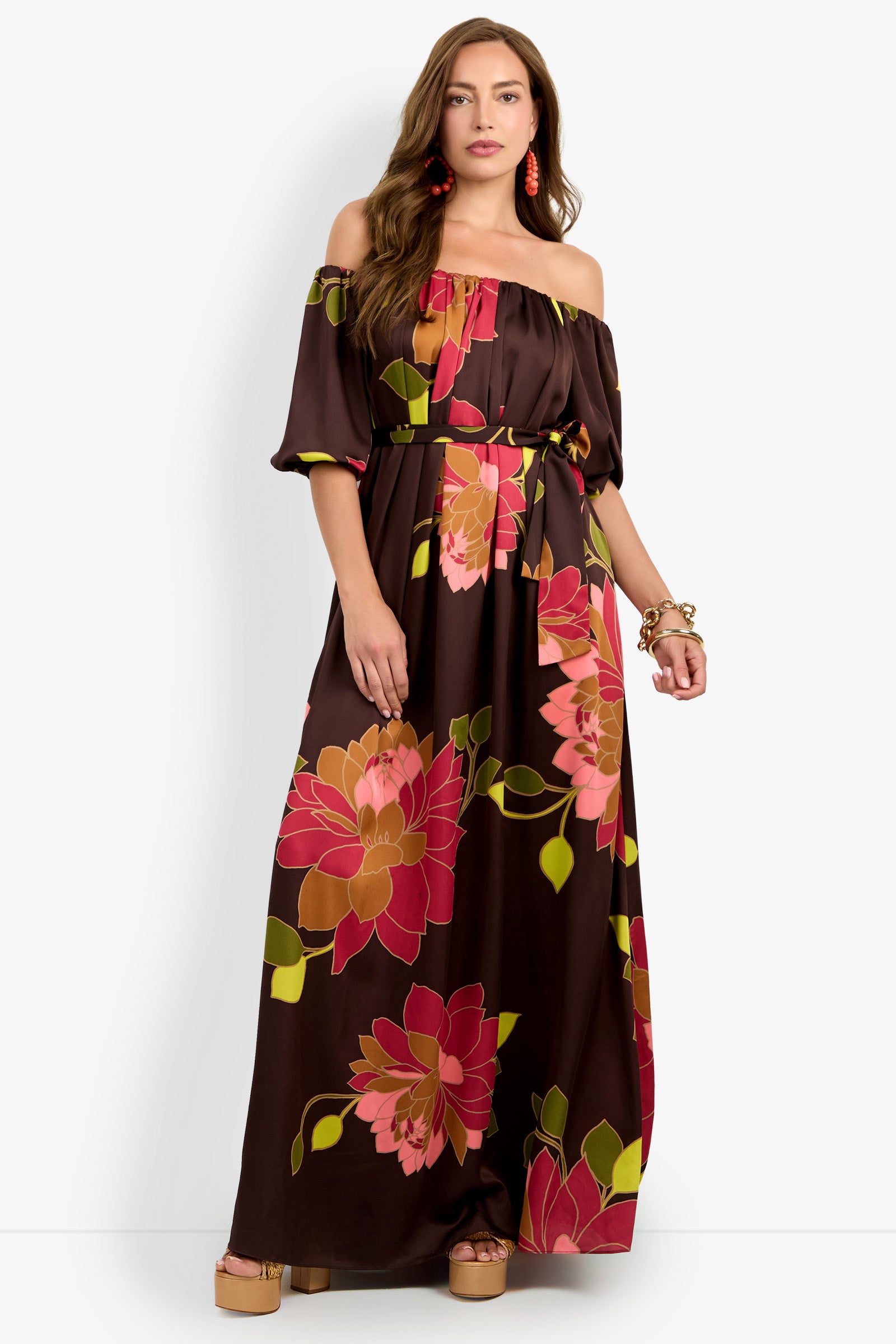 A woman stands against a white background wearing the Voglia Dress, a brown off-the-shoulder maxi dress featuring large pink and green floral prints, paired with tan platform sandals, red earrings, and bracelets.