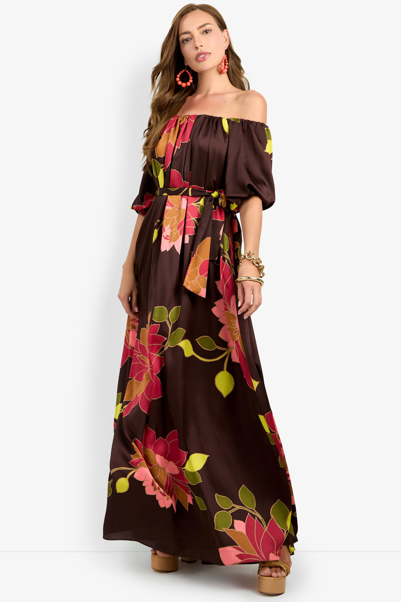 A woman wearing the Voglia Dress—an off-the-shoulder, brown maxi dress adorned with large pink and green floral prints—paired with tan platform sandals, orange statement earrings, and a bracelet, stands against a plain background.