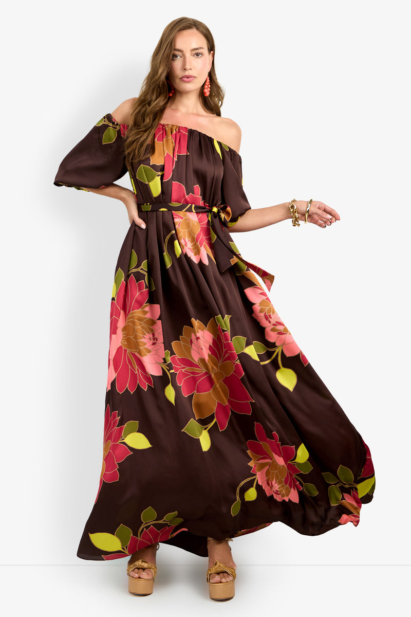 A woman stands against a plain background wearing the Voglia Dress, a flowing off-shoulder brown maxi adorned with large pink and green floral prints. She completes her look with tan platform sandals, gold jewelry, and pink earrings.