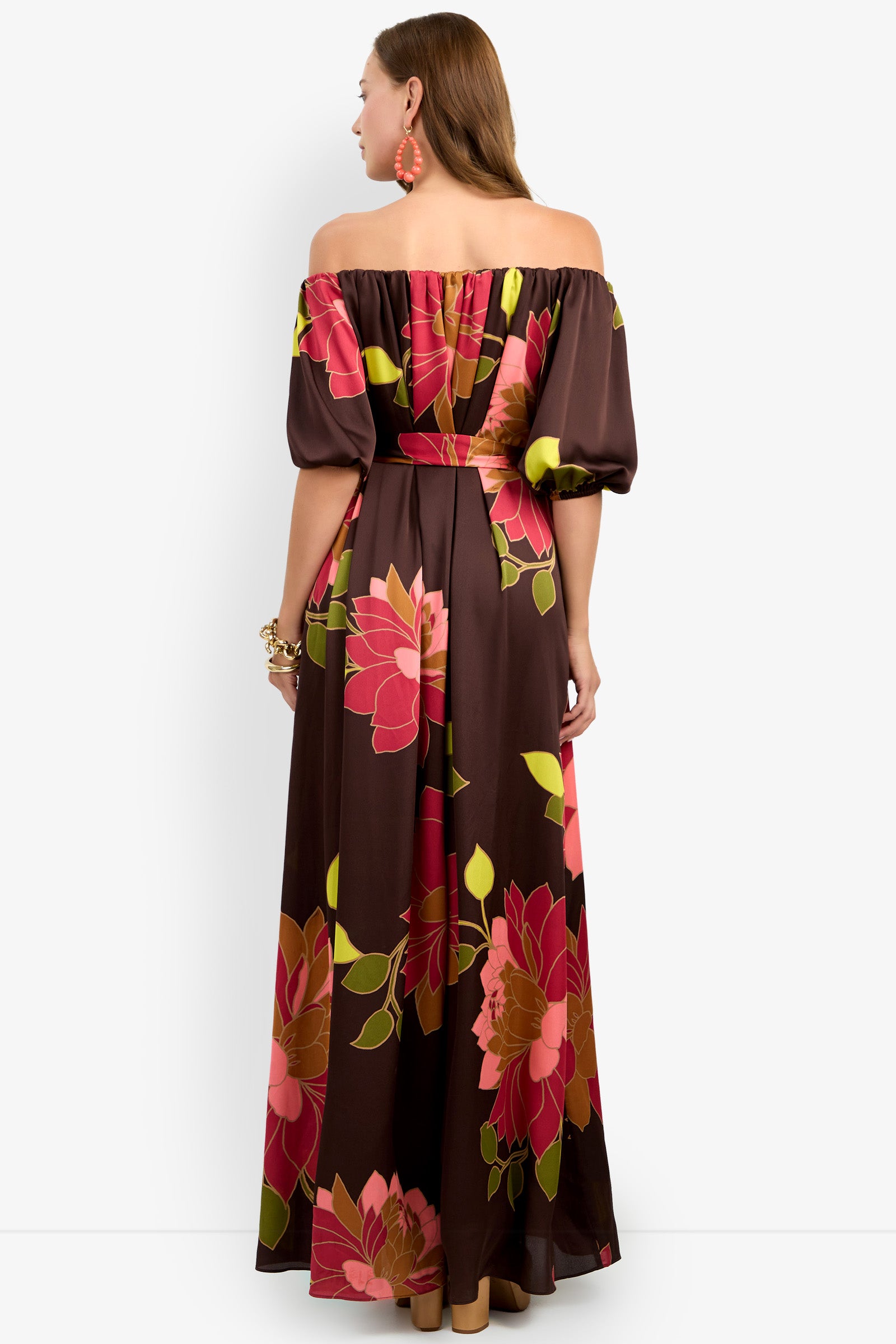 A woman facing away is wearing the Voglia Dress, a brown off-the-shoulder maxi dress featuring large pink, red, and yellow floral patterns. She has long hair, orange earrings, and a bracelet. The background is plain white.