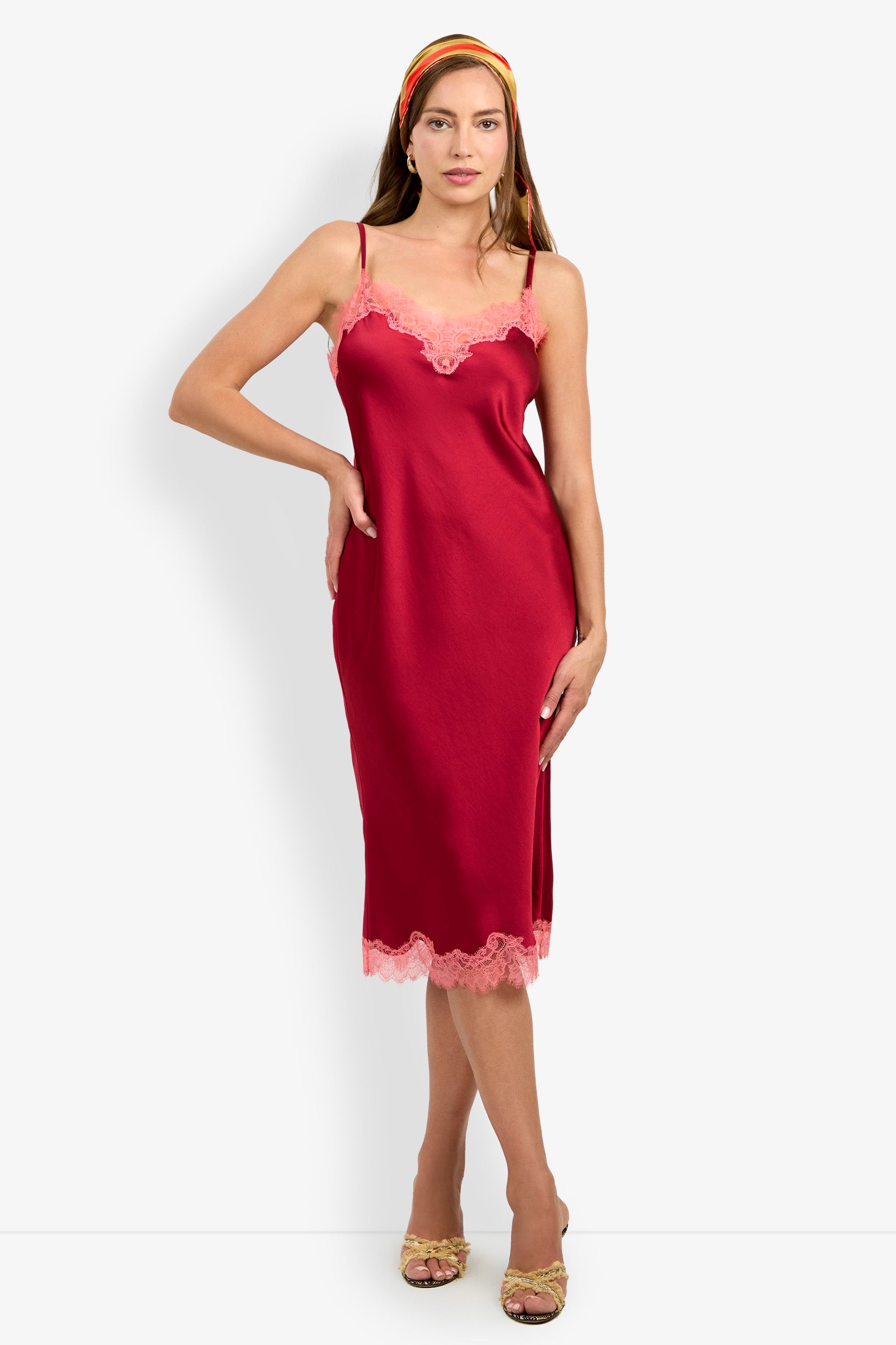A woman stands against a plain white background wearing the CARAMIA DRESS—a red satin slip dress with pink lace trim, crafted from 100% polyester—paired with a multicolored headband and strappy heeled sandals.