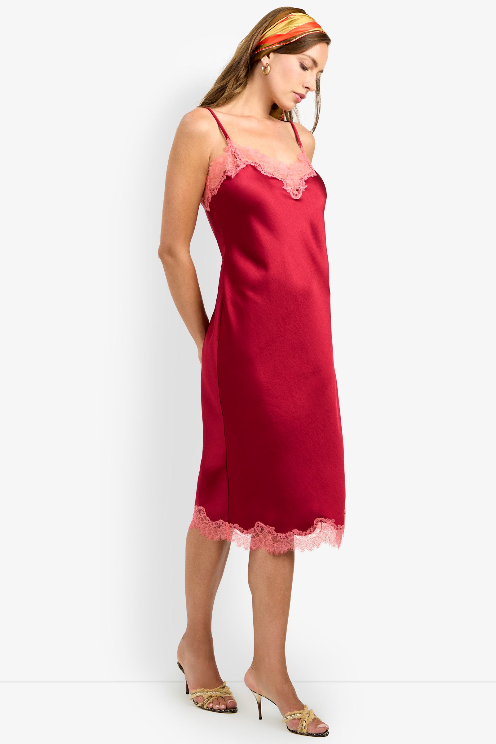 Woman wearing the CARAMIA DRESS, a red satin slip dress made from 100% polyester with lace trim, styled with gold heels and a colorful headscarf, standing against a plain white background.
