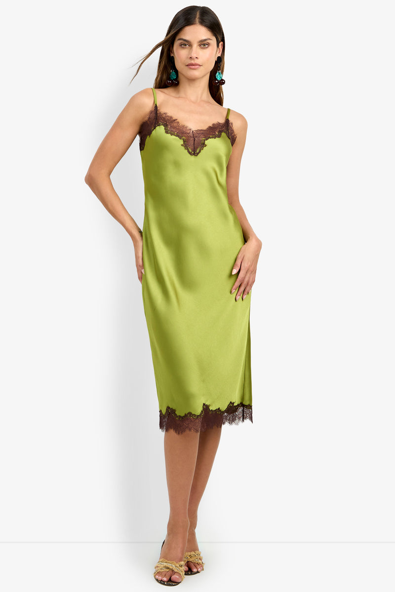 Wearing the CARAMIA DRESS—a light green satin slip dress made from luxurious 100% polyester, detailed with thin straps and dark lace trim at the neckline and hem—a woman pairs it with green earrings and open-toe heels as she stands against a plain white background.