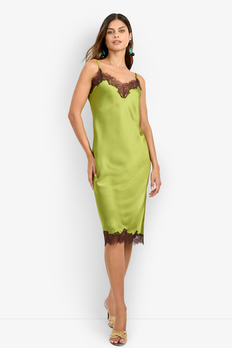 Wearing the CARAMIA DRESS—a sleeveless, lime green satin slip crafted from 100% polyester and detailed with black lace trim at the neckline and hem—a woman pairs it with open-toe heels and long earrings as she poses against a plain white background.