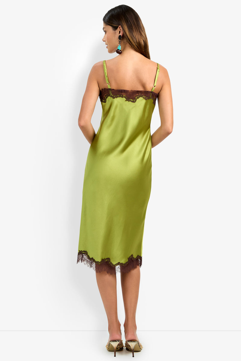 Wearing the CARAMIA DRESS—crafted from luxurious green satin polyester with elegant black lace trim—a woman stands with her back to the camera. Her long brown hair cascades down, complemented by heels and statement earrings, all set against a plain white background.