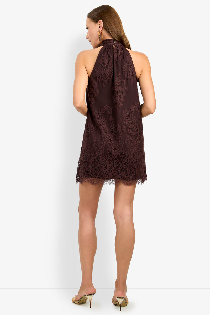 A woman stands facing away against a plain white background, wearing the ZANA DRESS—a sleeveless dark lace halter dress with a short hem—paired with gold high heels.