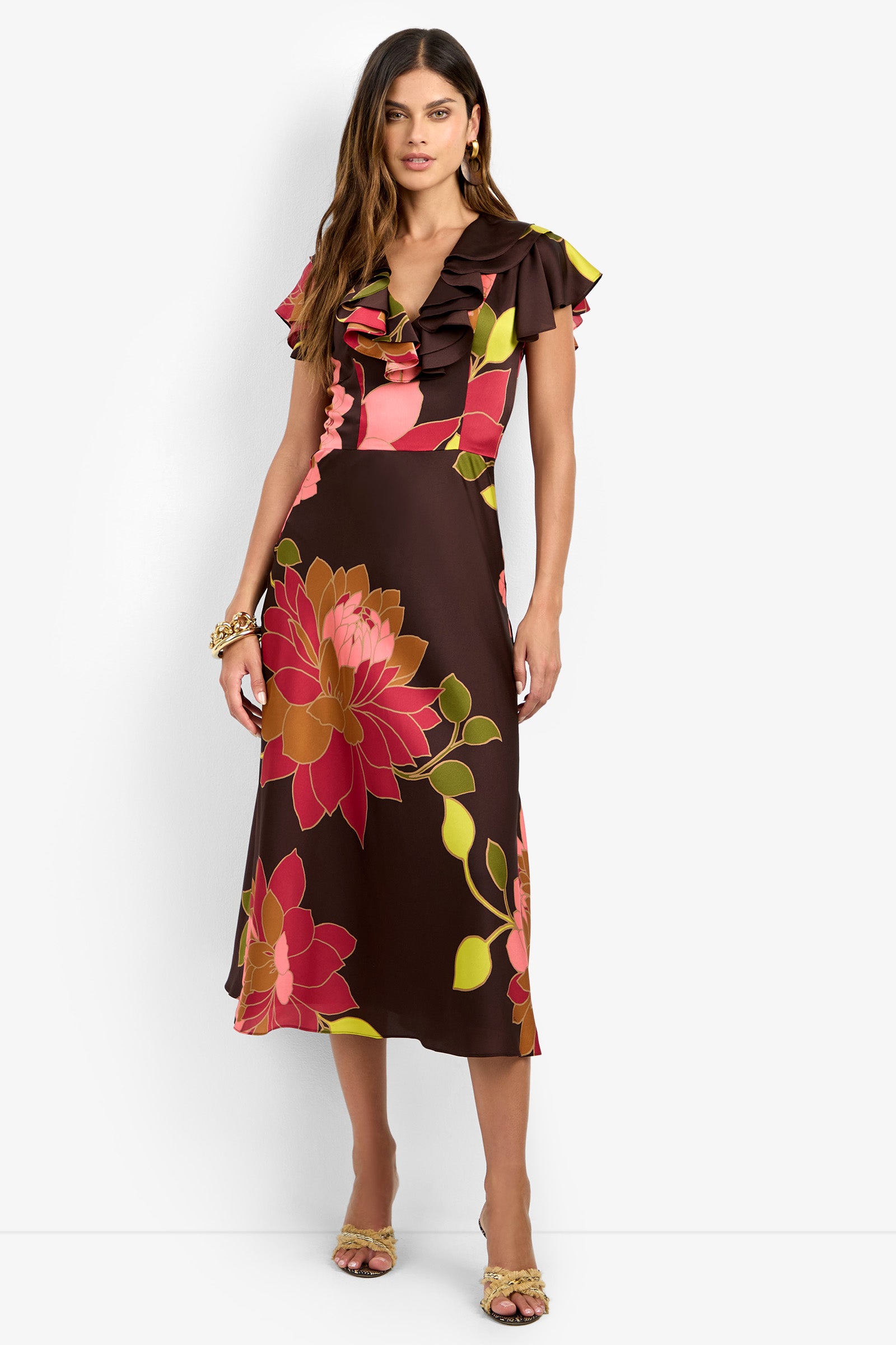 Woman wearing the PATRONS DRESS—a brown, pink, and yellow floral midi dress with ruffled sleeves—styled with a gold bracelet and yellow heeled sandals, standing against a white background.