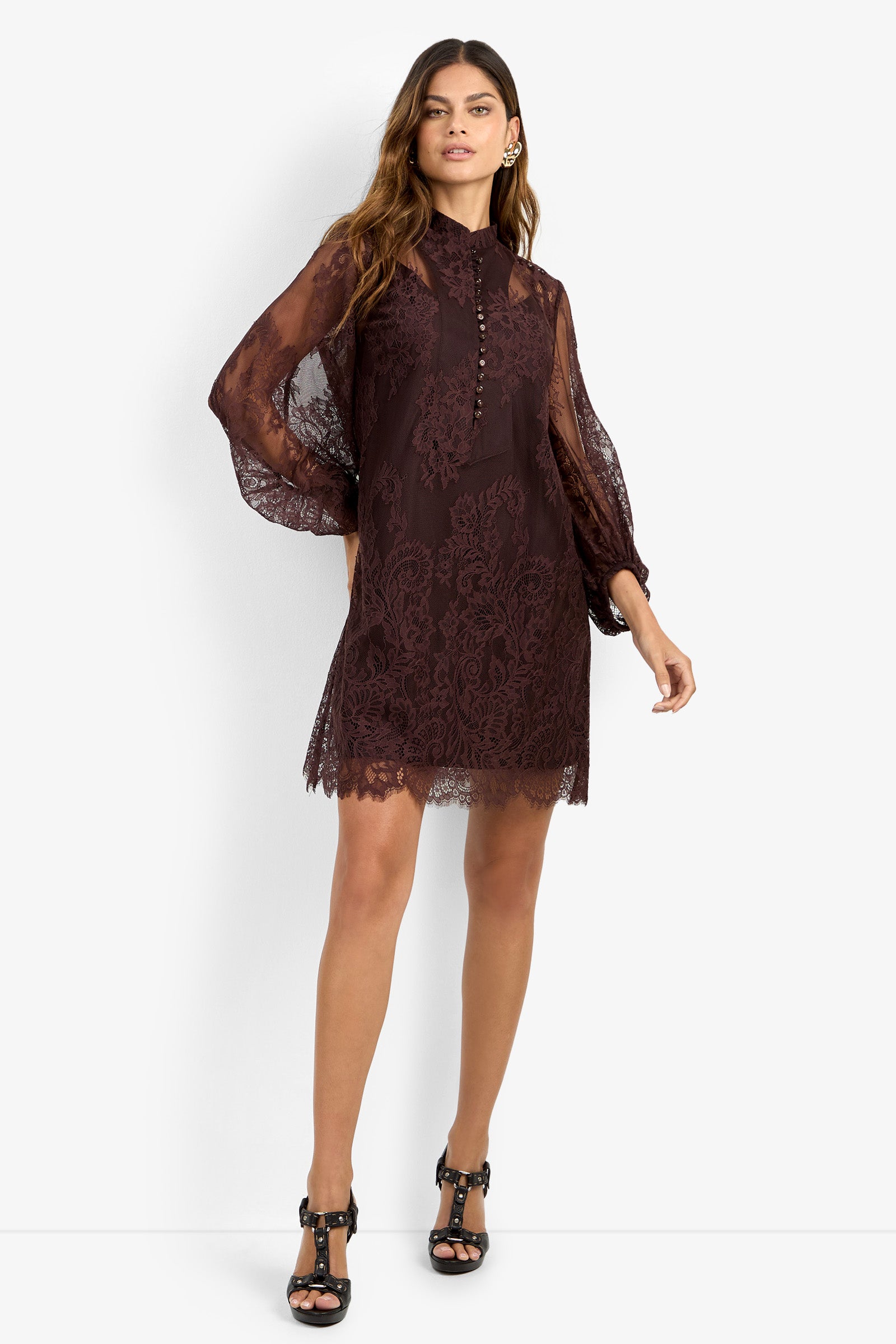 A woman wearing the BOHEME DRESS—a dark lace mini dress with sheer long sleeves and a buttoned high neck—paired with black heels, stands against a plain white background.