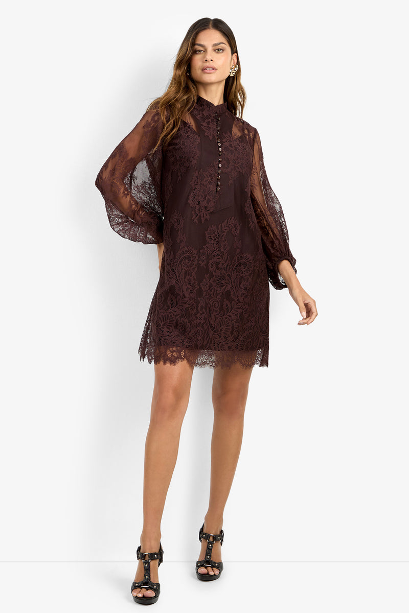 A woman wearing the BOHEME DRESS—a dark lace mini dress with sheer long sleeves and a buttoned high neck—paired with black heels, stands against a plain white background.