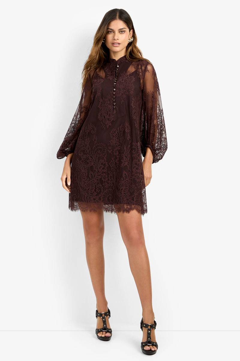 A woman stands against a plain white background wearing the BOHEME DRESS—featuring sheer, long sleeves, dark lace fabric, and front buttons—and black heeled sandals, with one hand resting relaxed by her side.