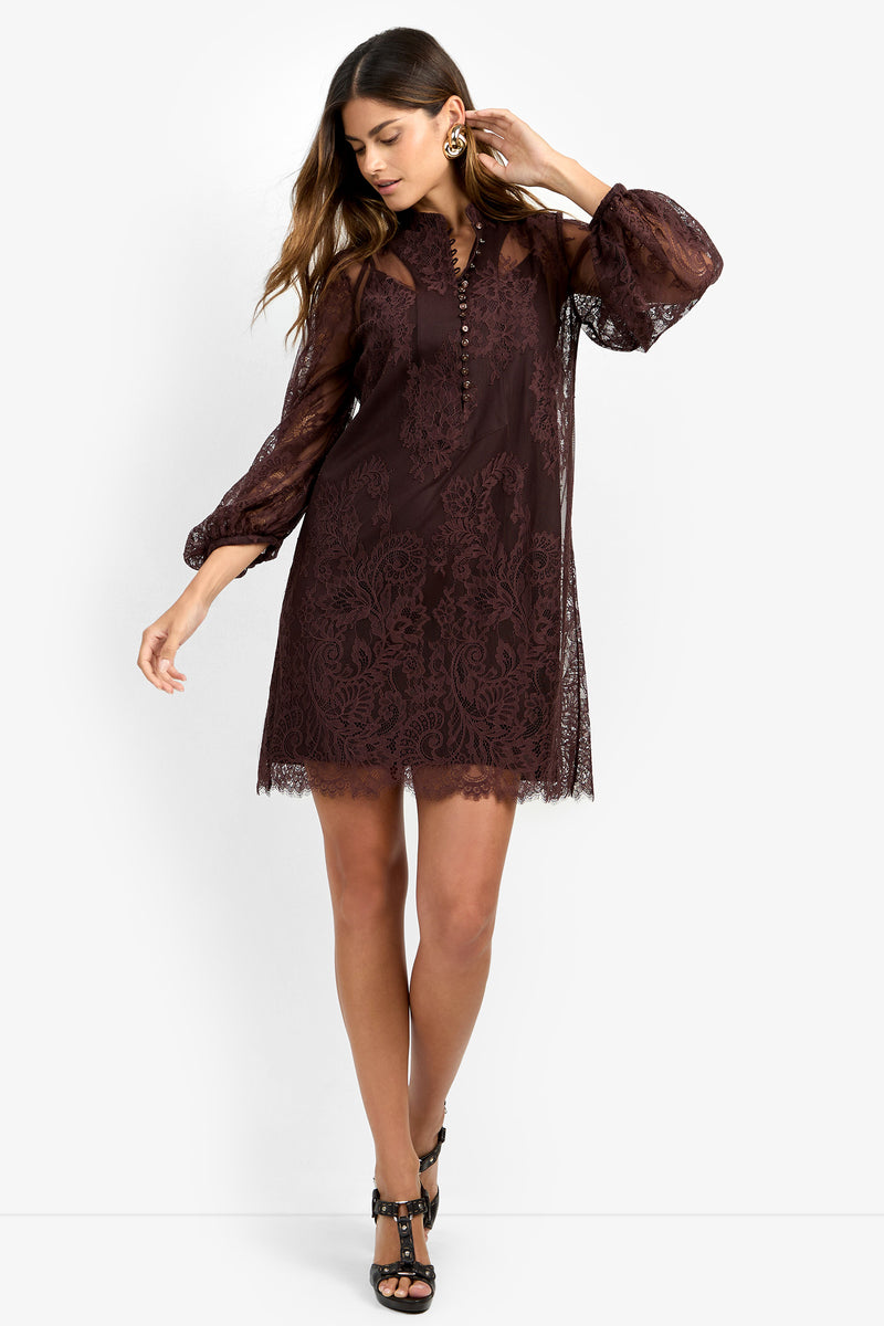 A woman models the BOHEME DRESS—a knee-length, long-sleeve burgundy lace dress featuring a high neck and button details—as she poses against a white background with one hand touching her earring and the other by her side.