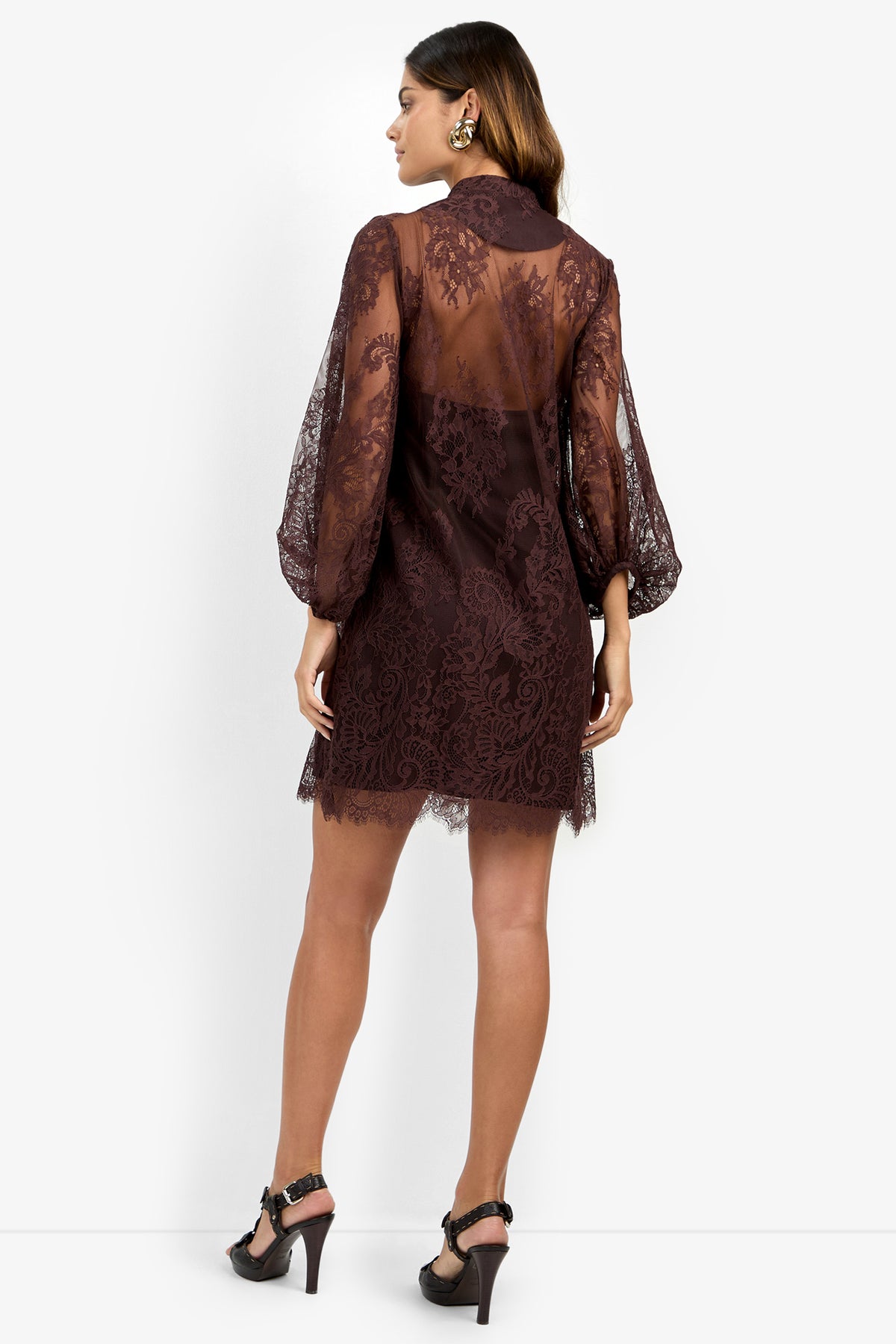 A woman stands with her back to the camera, wearing the BOHEME DRESS—a sheer, long-sleeved, dark lace design—paired with high-heeled sandals. Her hair is down, and she accessorizes with earrings.