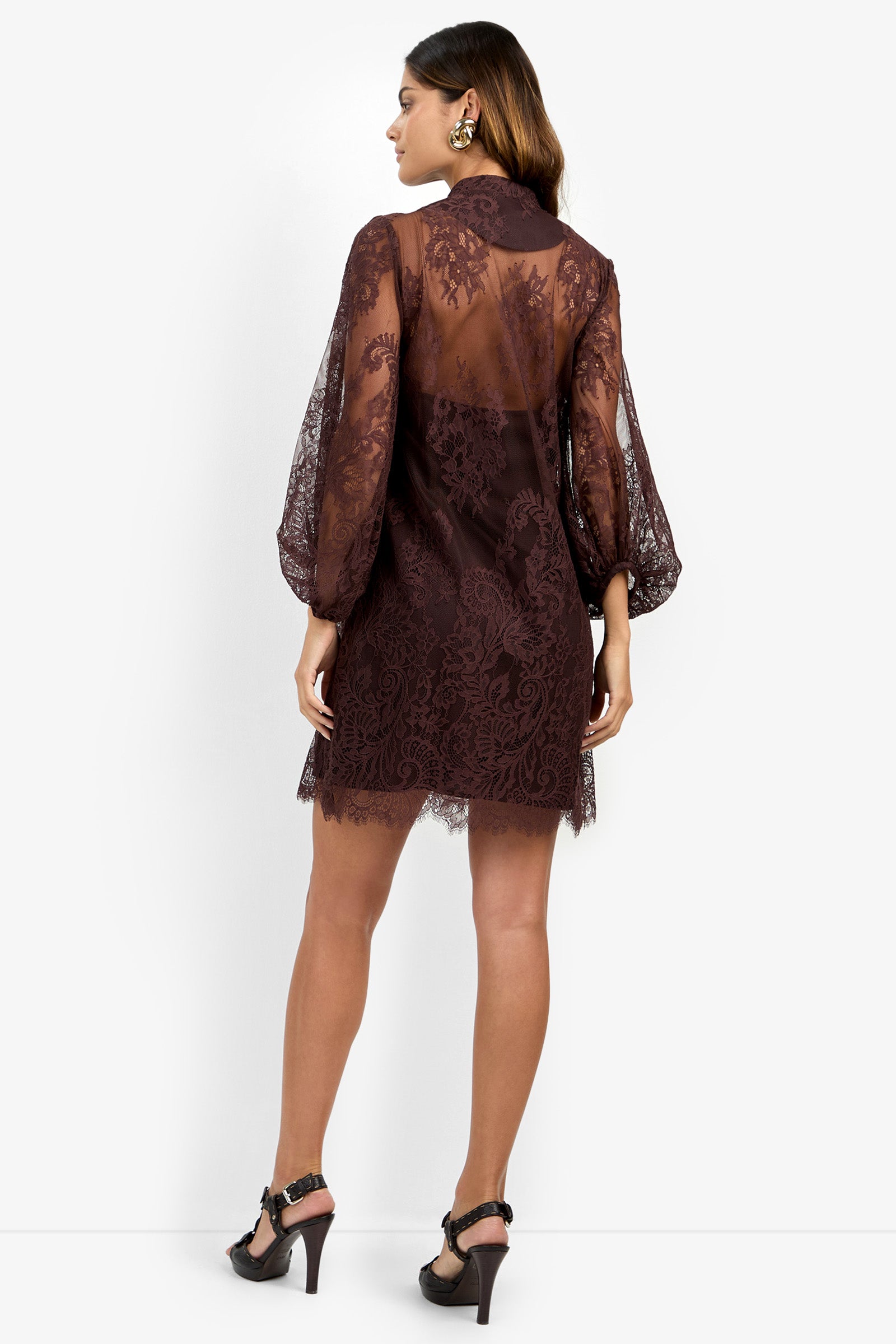 A woman stands with her back to the camera, wearing the BOHEME DRESS—a sheer, long-sleeved, dark lace design—paired with high-heeled sandals. Her hair is down, and she accessorizes with earrings.