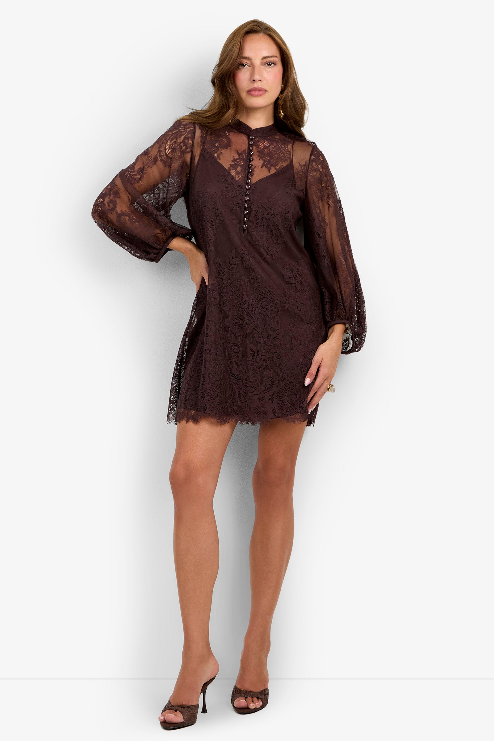 A woman poses against a plain white background, wearing the Boheme Dress—a dark lace mini dress featuring long sheer sleeves and button details—paired with open-toe heels.
