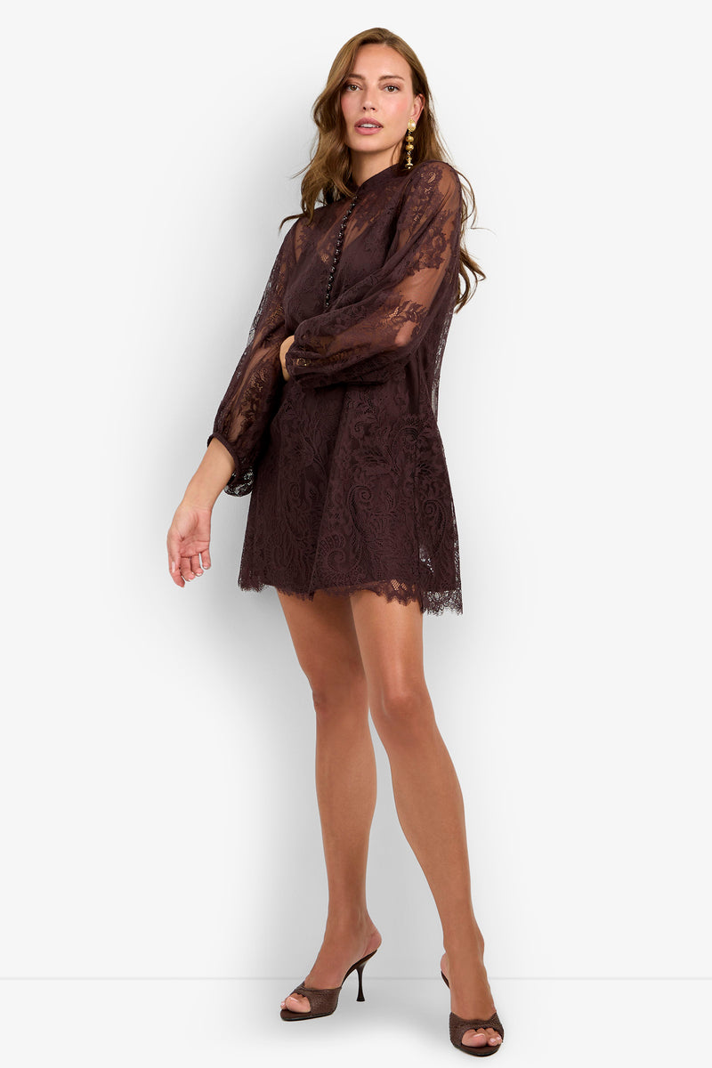 A woman stands against a plain white background, one arm bent, wearing the Boheme Dress—a sheer, long-sleeve dark lace mini with a high neckline and scalloped hem—paired with dark open-toe heels.