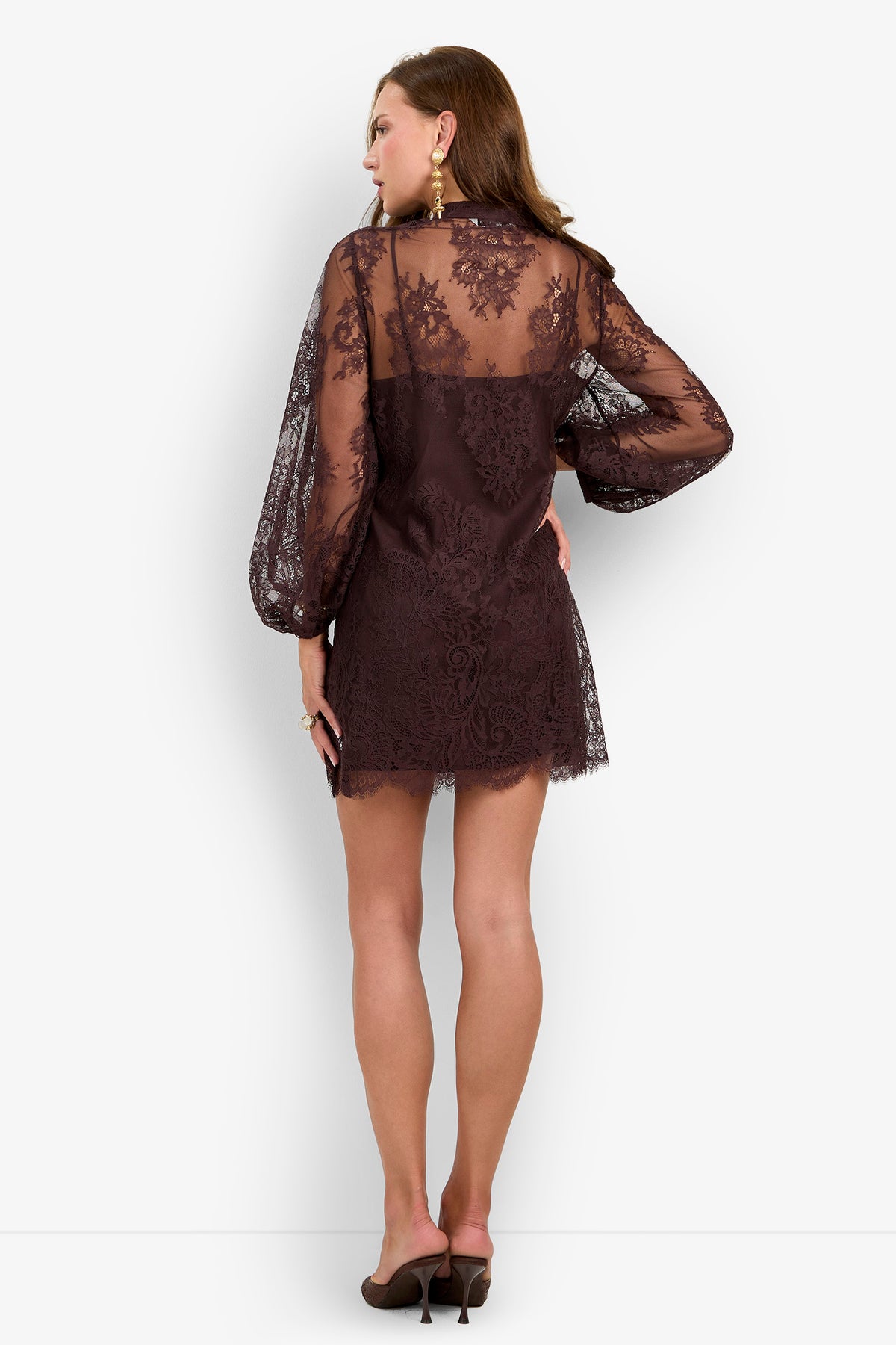 A woman stands facing away, showcasing the back details of the Boheme Dress—a sheer, dark lace design with long sleeves—paired with high heels.