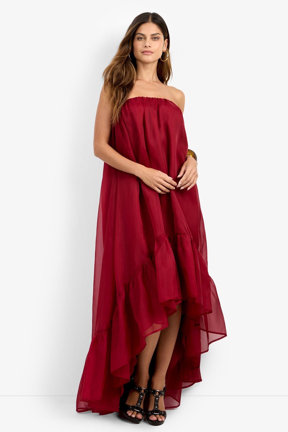 A woman wearing the ARIAH DRESS, a strapless, flowy high-low burgundy dress with a ruffle hem, stands against a white background. She has long hair, gold hoop earrings, and black strappy heels.
