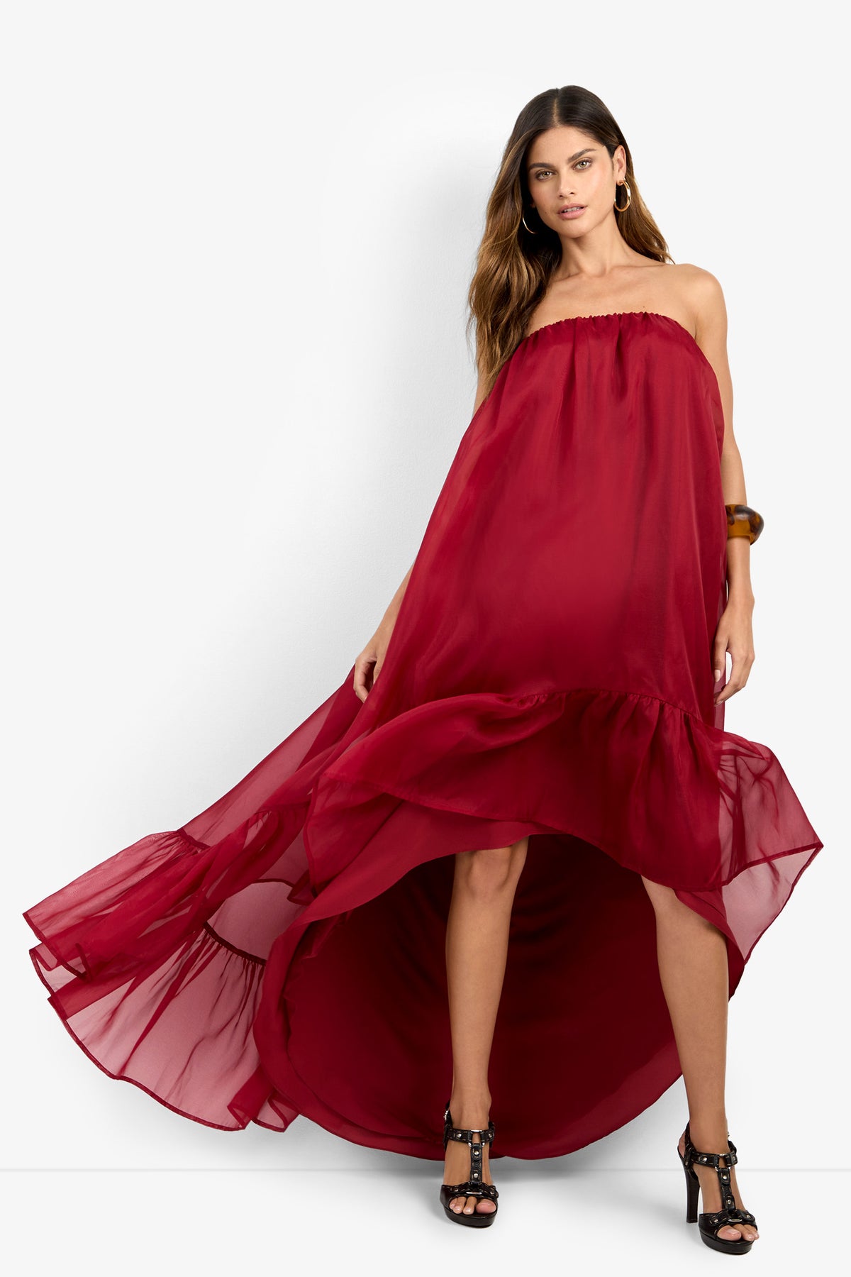 A woman stands confidently with one hand on her hip, wearing the ARIAH DRESS—a strapless, flowing red high-low dress featuring a ruffled hem that billows outward. She completes the look with black heels and a brown bracelet.