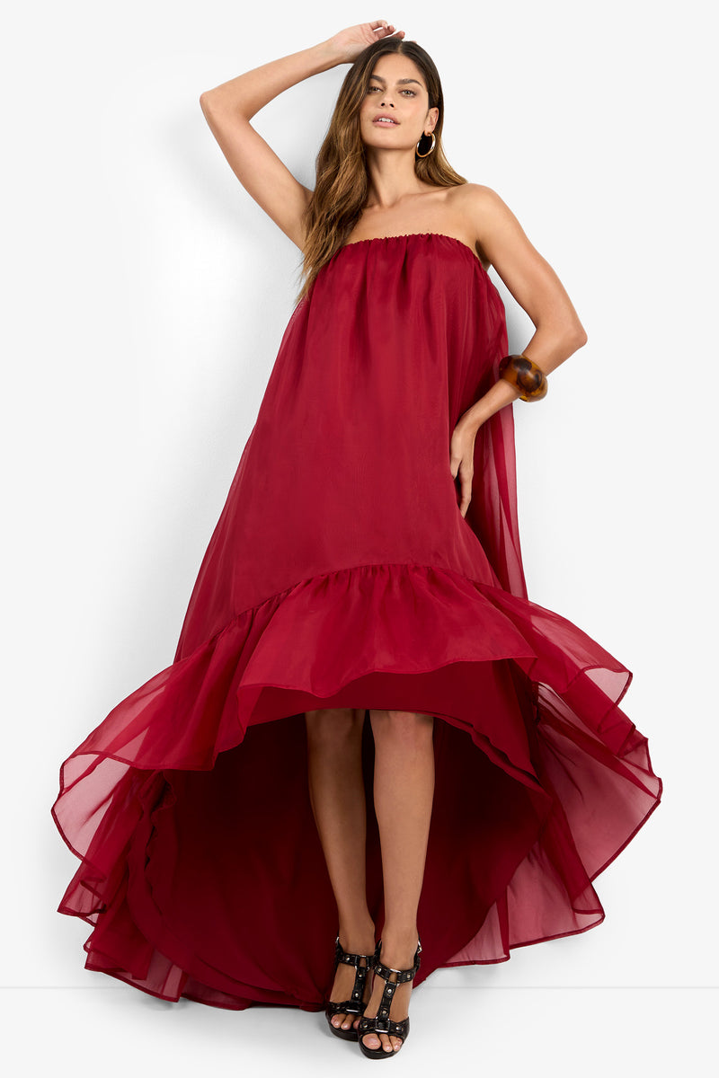 A woman in the ARIAH DRESS — a strapless, high-low red gown with a flowing silhouette and ruffled hem — styled with black strappy heels, gold hoop earrings, and a chunky brown bracelet, stands with one arm raised and the other holding her dress.