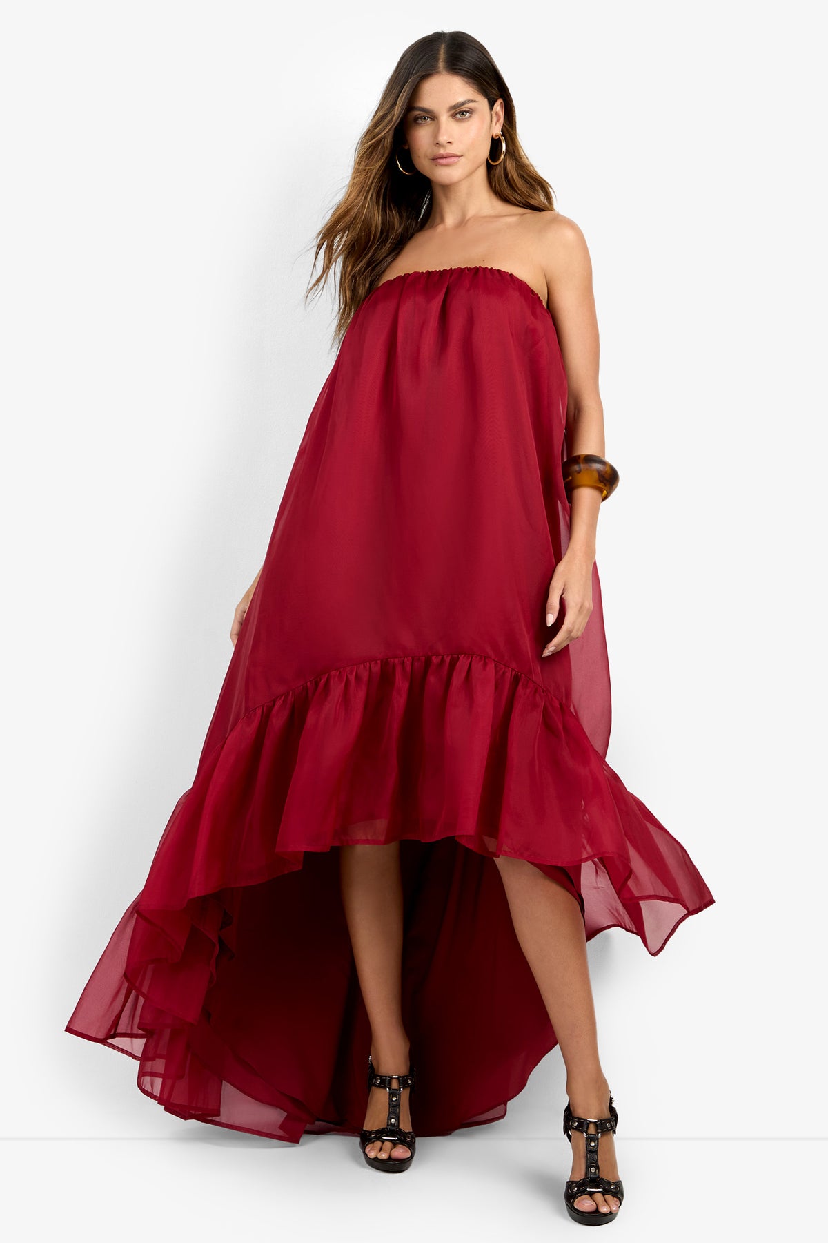 A woman wears the ARIAH DRESS, a strapless, high-low burgundy dress with a ruffled hem, paired with black heels and a brown bangle. Her long hair is styled loose as she stands against a plain white background.