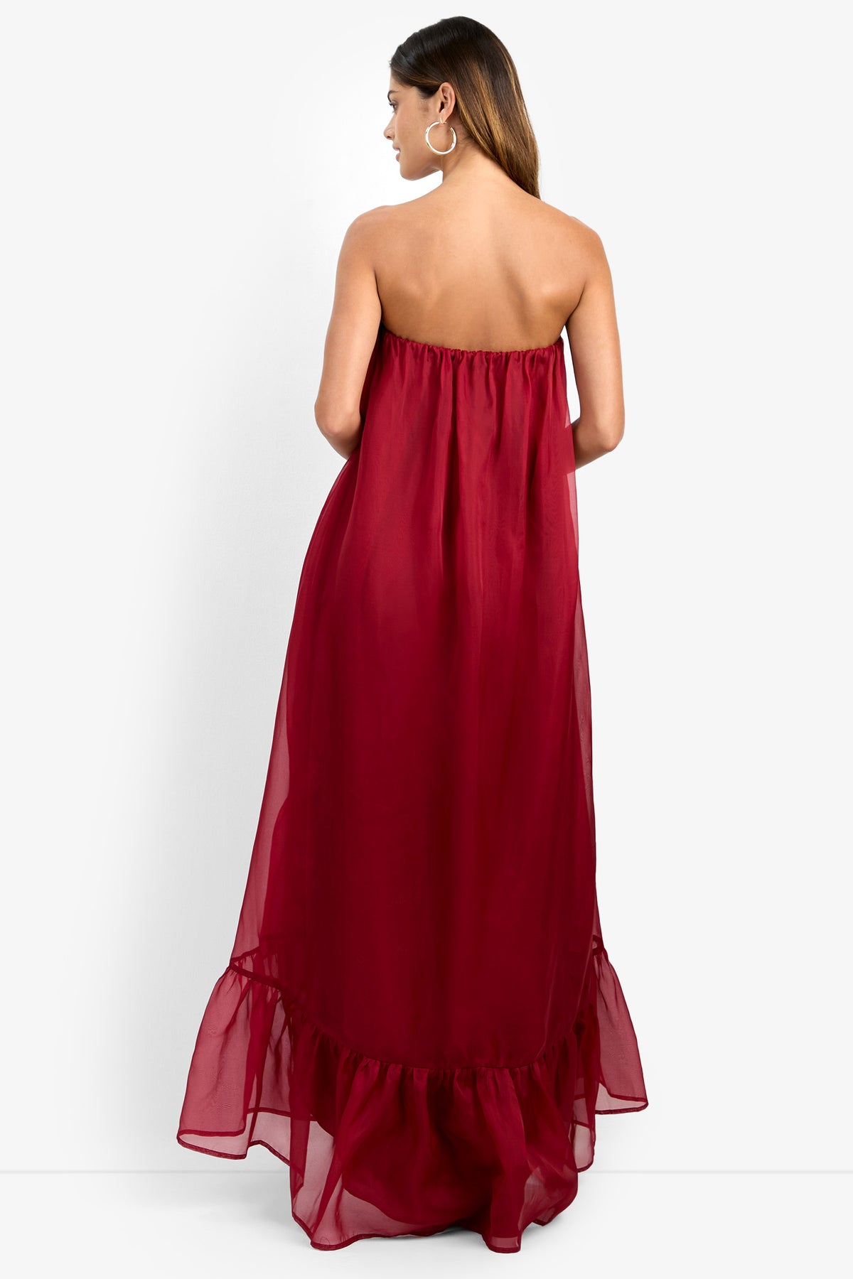 A woman stands facing away, wearing the ARIAH DRESS—a strapless, floor-length burgundy gown featuring a ruffled hem. She accessorizes with large hoop earrings, and her long hair is swept to one side against a plain white background.
